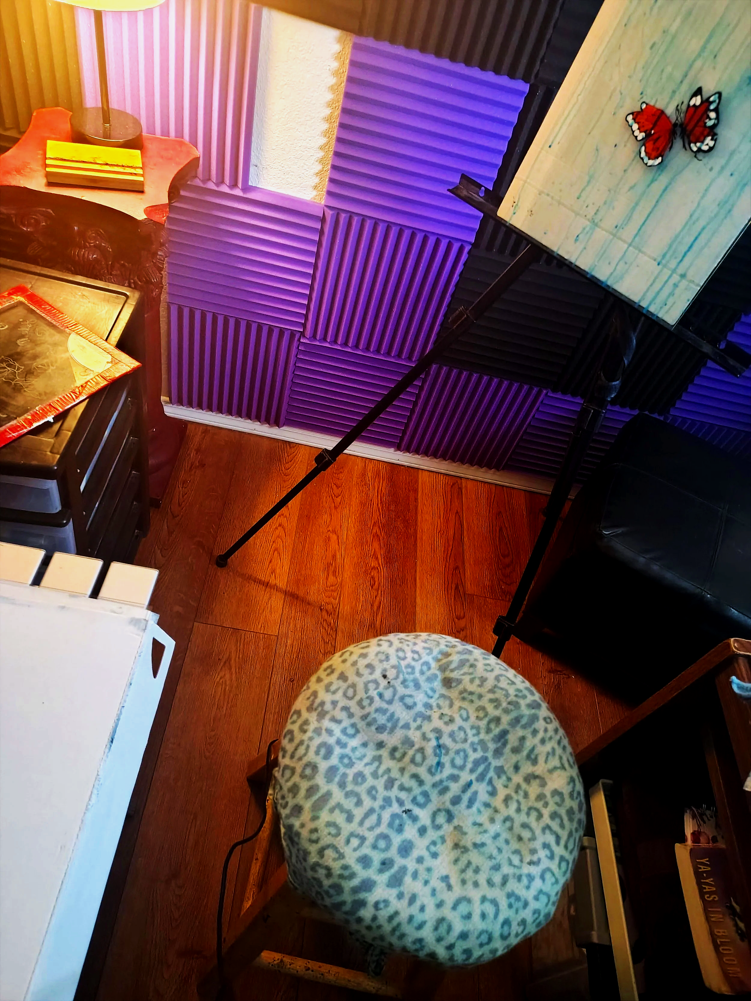 A room setup with purple and black soundproofing foam on the walls. There is a small table with a butterfly decoration on it, a black leather chair, and a wooden chair with a leopard print cushion in the foreground. The room has wooden flooring.