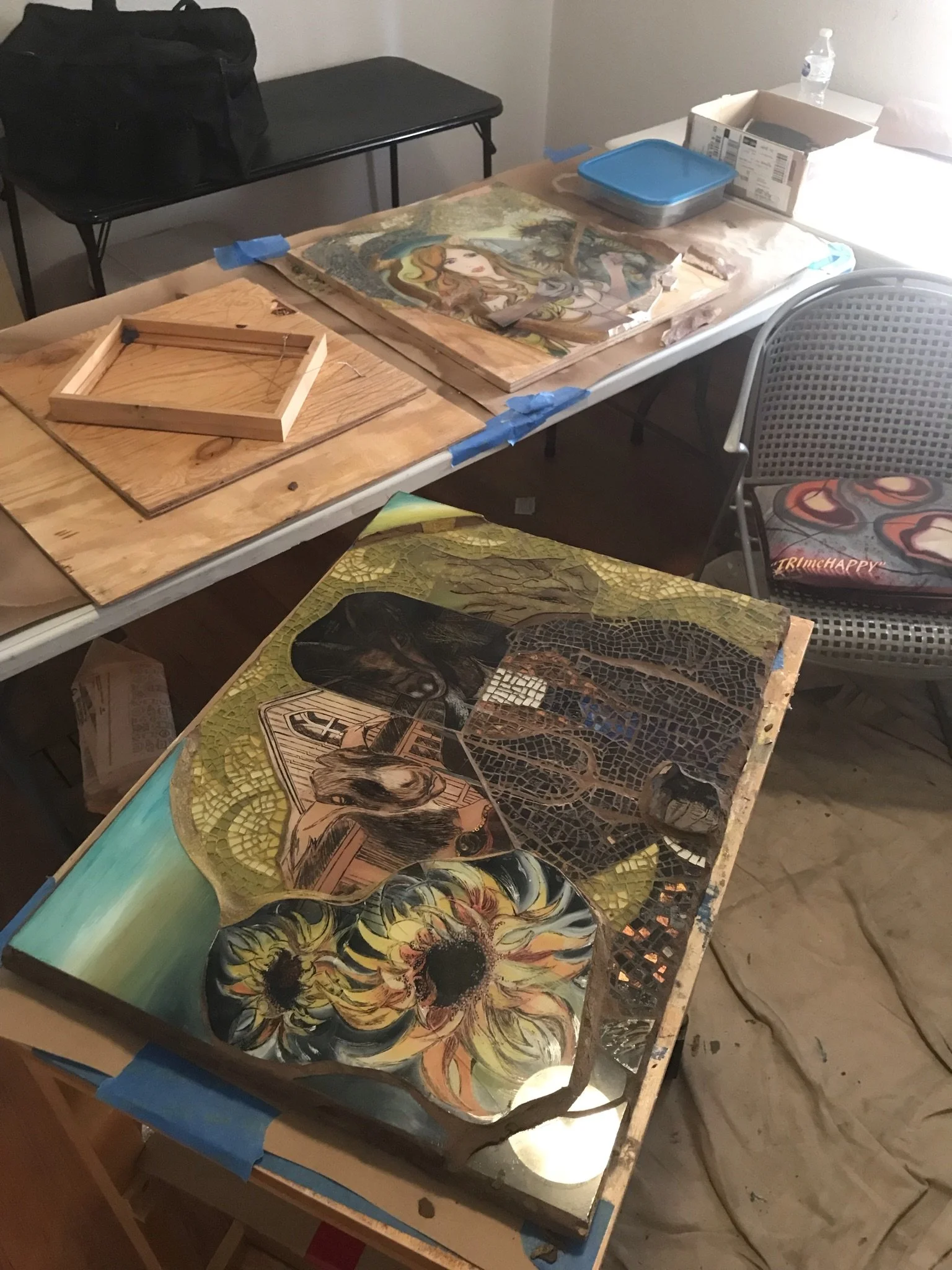A workspace with various unfinished art projects, including painted and mosaic pieces, on tables. There are art supplies, a water bottle, and a chair with a colorful cushion.