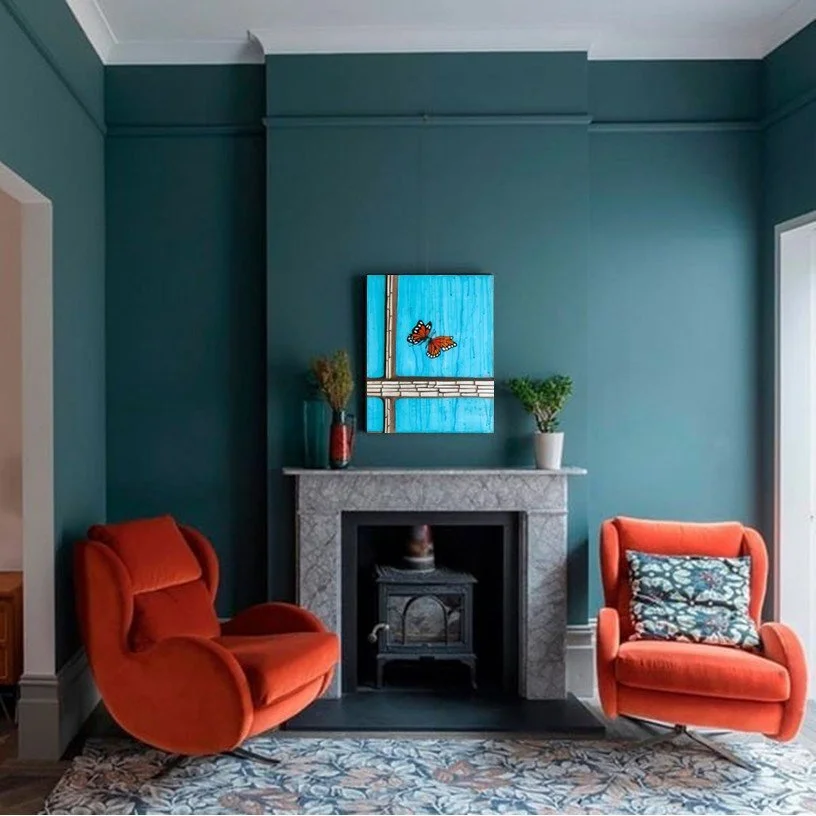 Living room corner with teal walls, a white marble fireplace, a small black stove inside the fireplace, two orange armchairs, one with a patterned cushion, and a floral area rug. There is a colorful painting of butterflies on the wall above the fireplace, and two potted plants on the mantel.