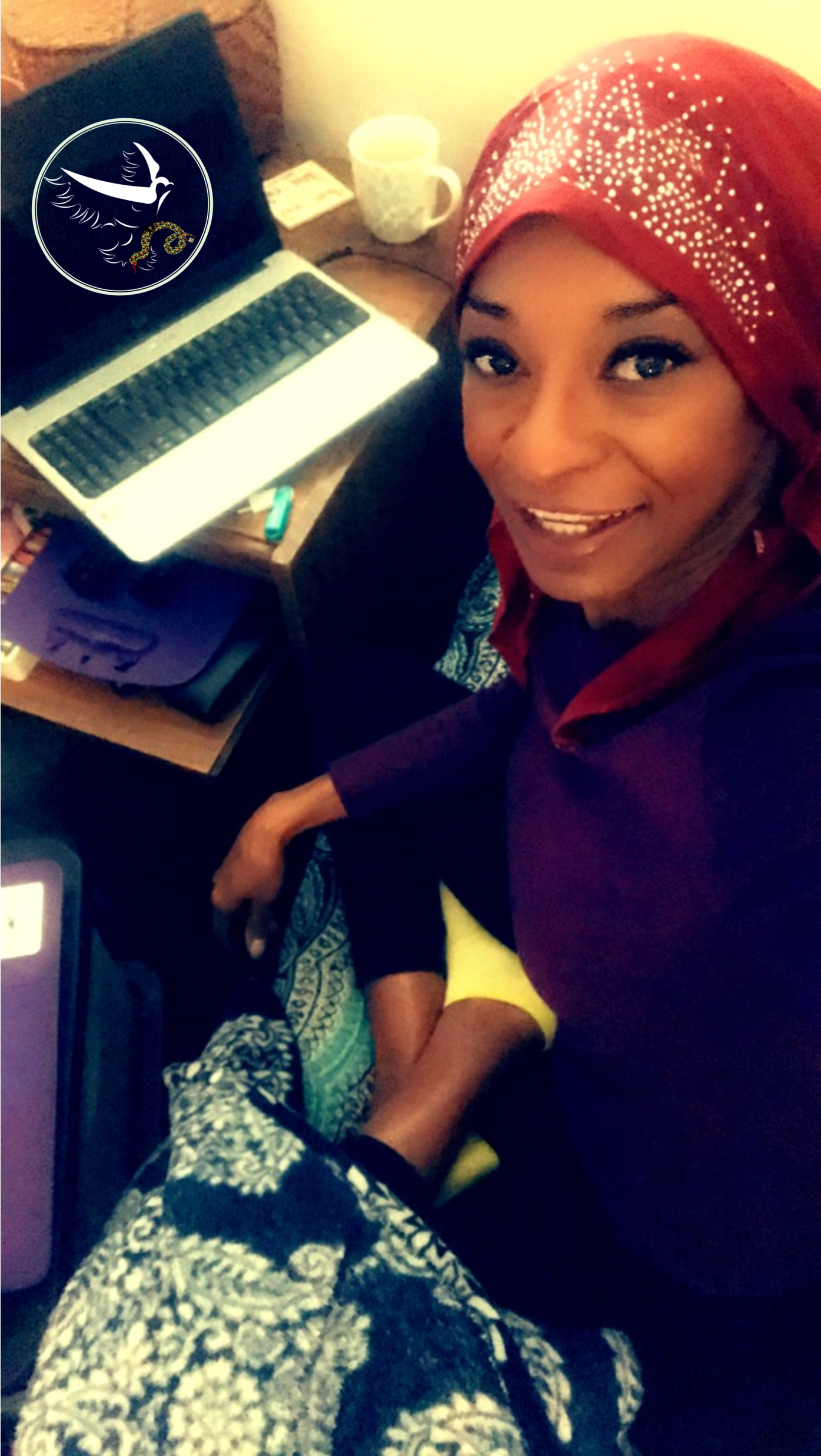 A woman wearing a red headscarf with rhinestones, smiling, sitting at a desk with a laptop, a mug, and a small box. She is wearing a maroon top with a yellow sleeve visible.