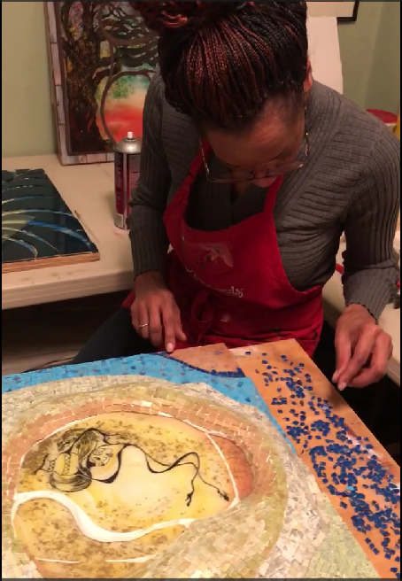 A woman with braided hair wearing glasses, a gray sweater, and a red apron, working on a mosaic art piece that features a snake.