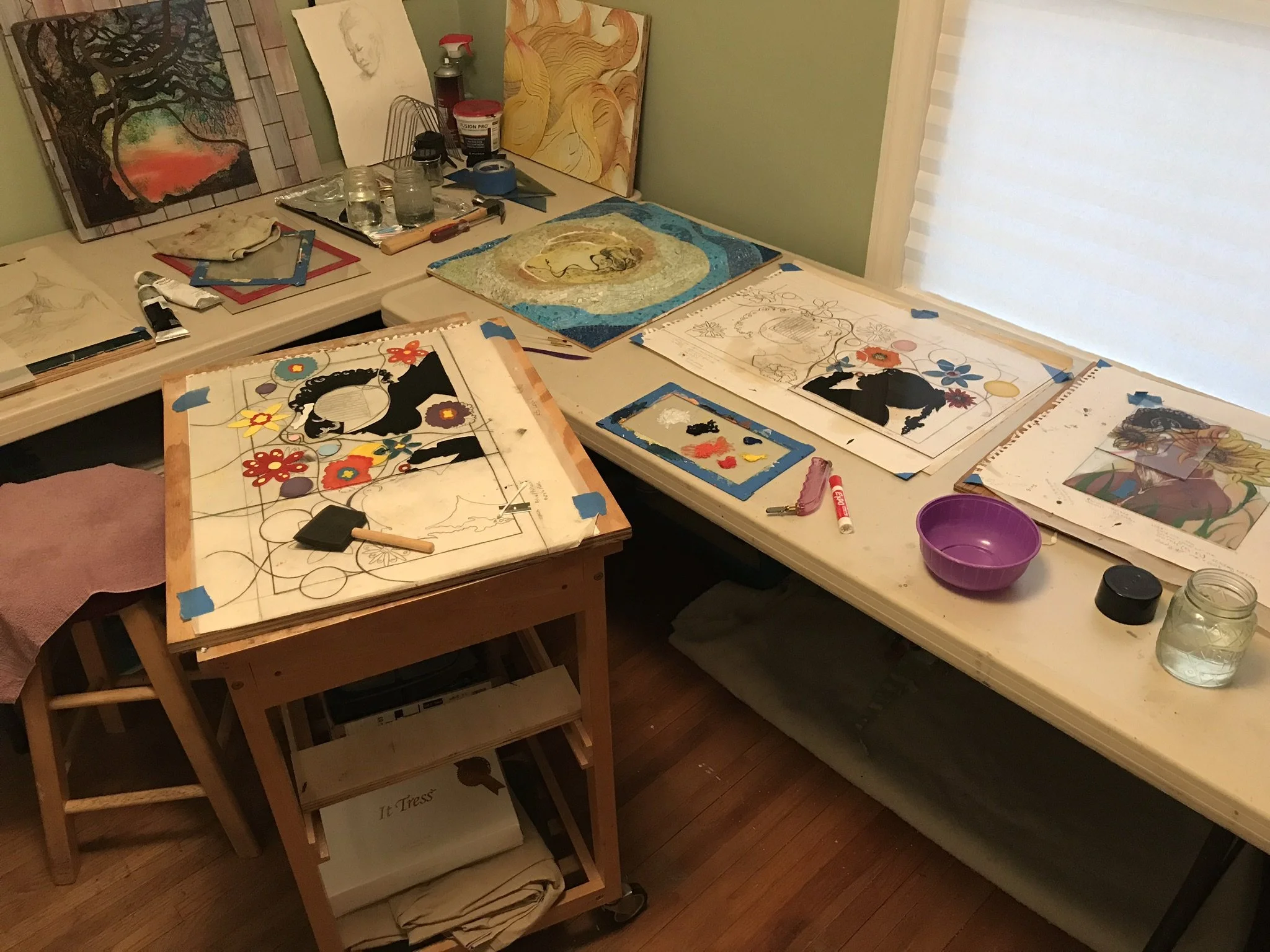 An art studio workspace with paintings and sketches on tables and walls, art supplies, Mason jars, and water jars on the table.