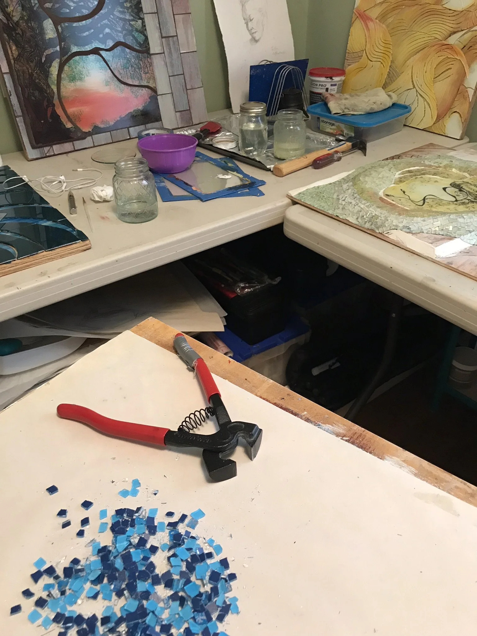 Artists' workspace with mosaic artwork in progress, mosaic tiles scattered on the table, and various art supplies including a hammer, pliers, jars of water, and completed or partially completed mosaics.