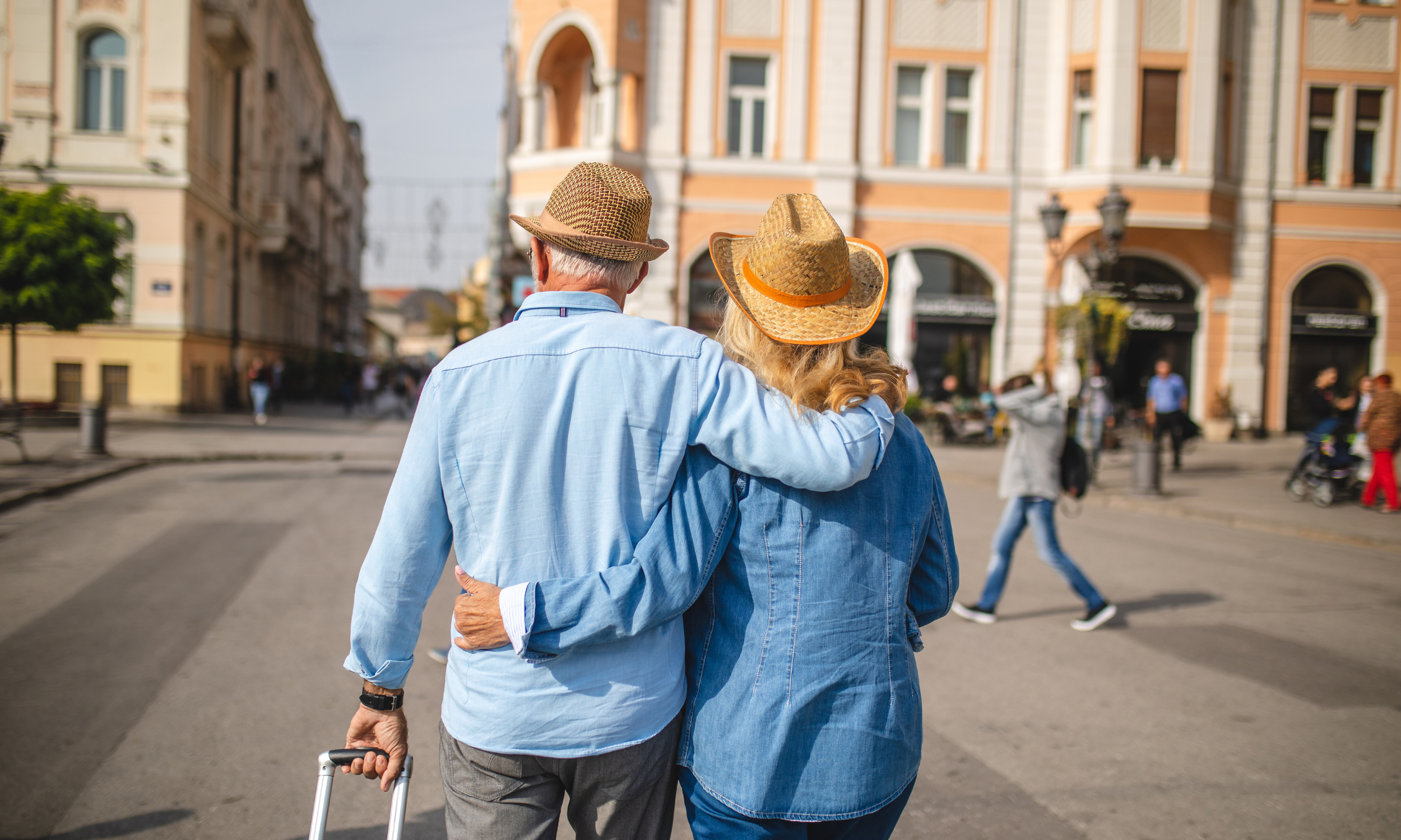 An elderly couple walking arm-in-arm through a city street, both wearing straw hats, with the man using a cane, back view.