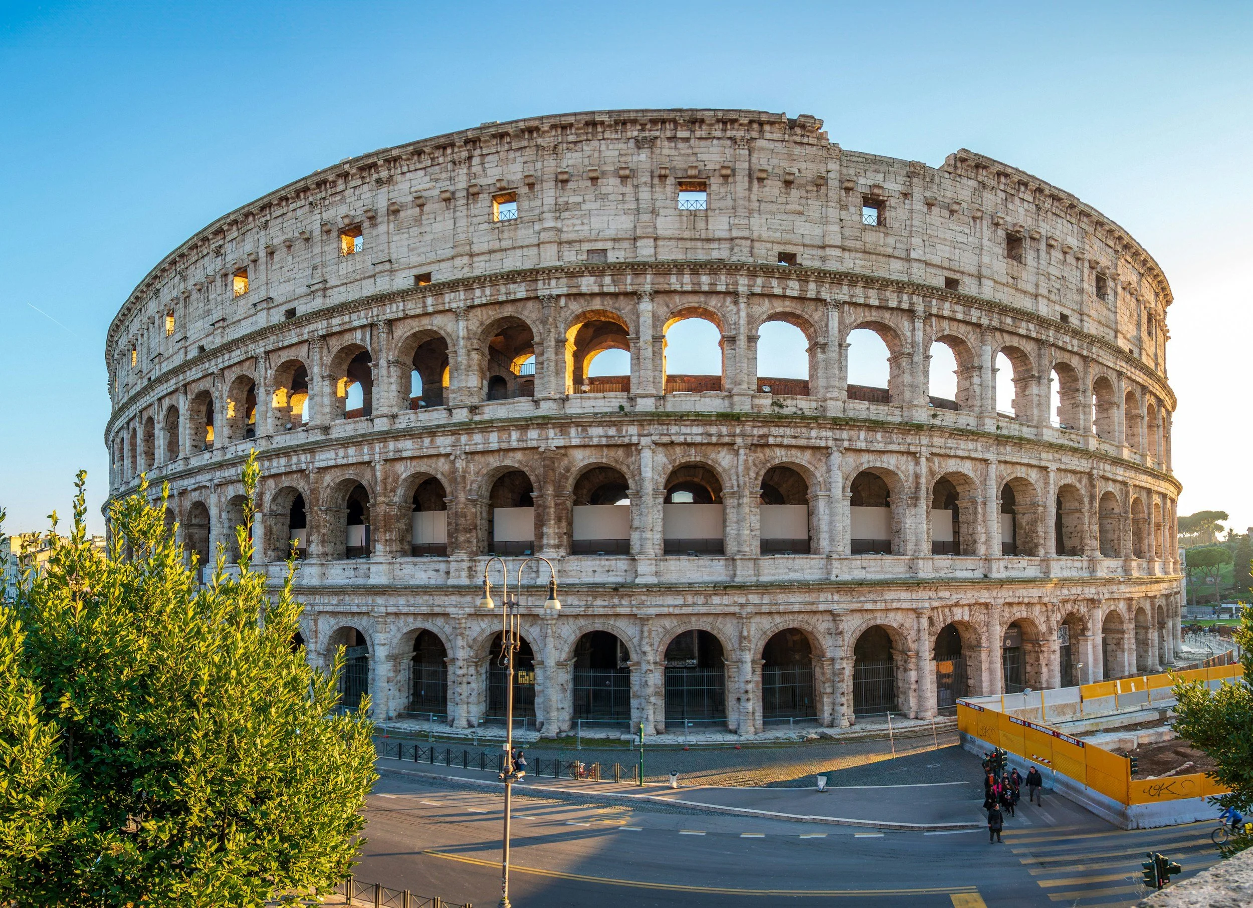 The ancient Roman Colosseum in Rome, Italy, with its stone arches and partially illuminated windows, shown during the day with blue sky.