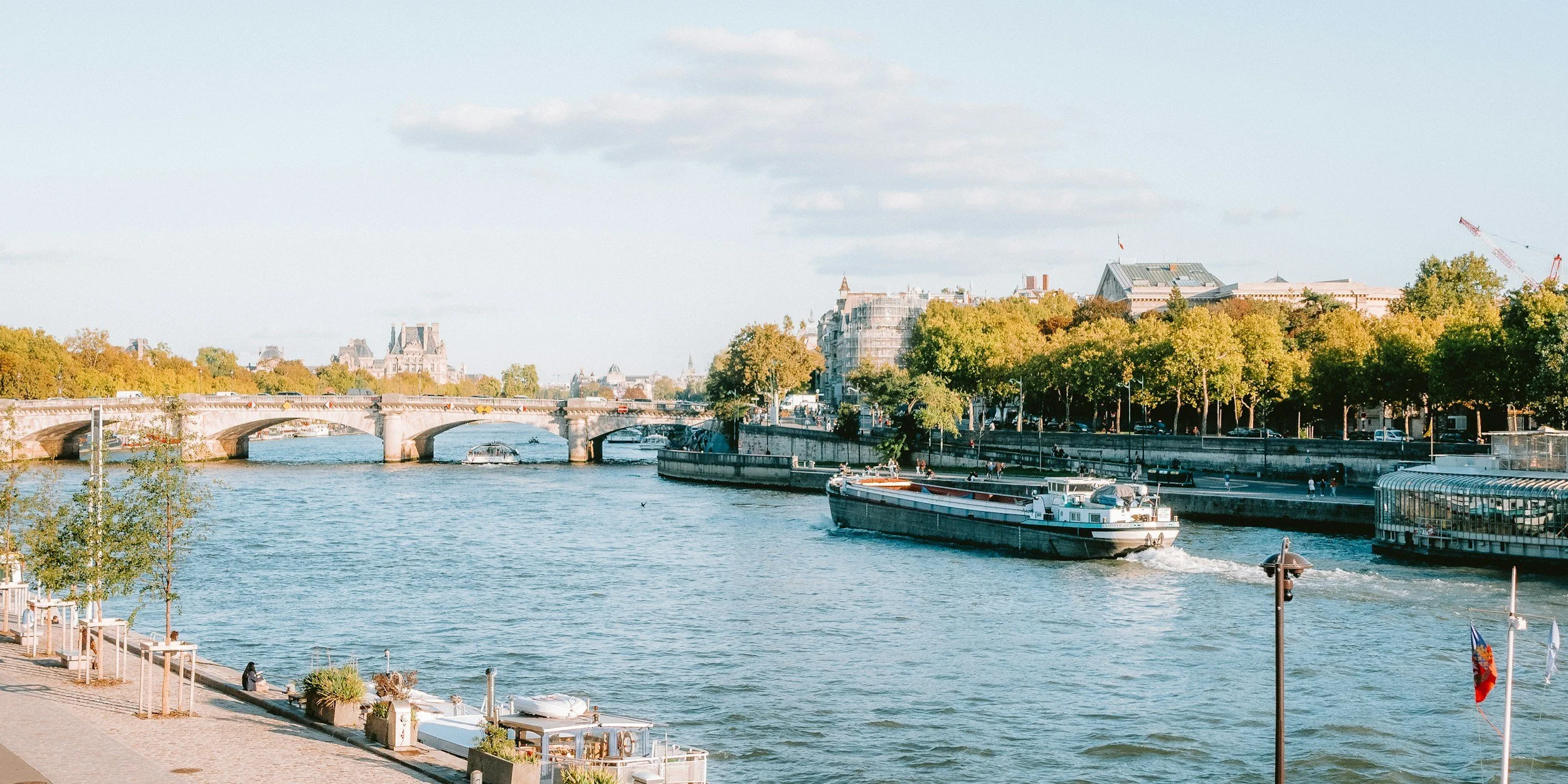 A river with a bridge, boats, and trees along the riverbank on a clear day.