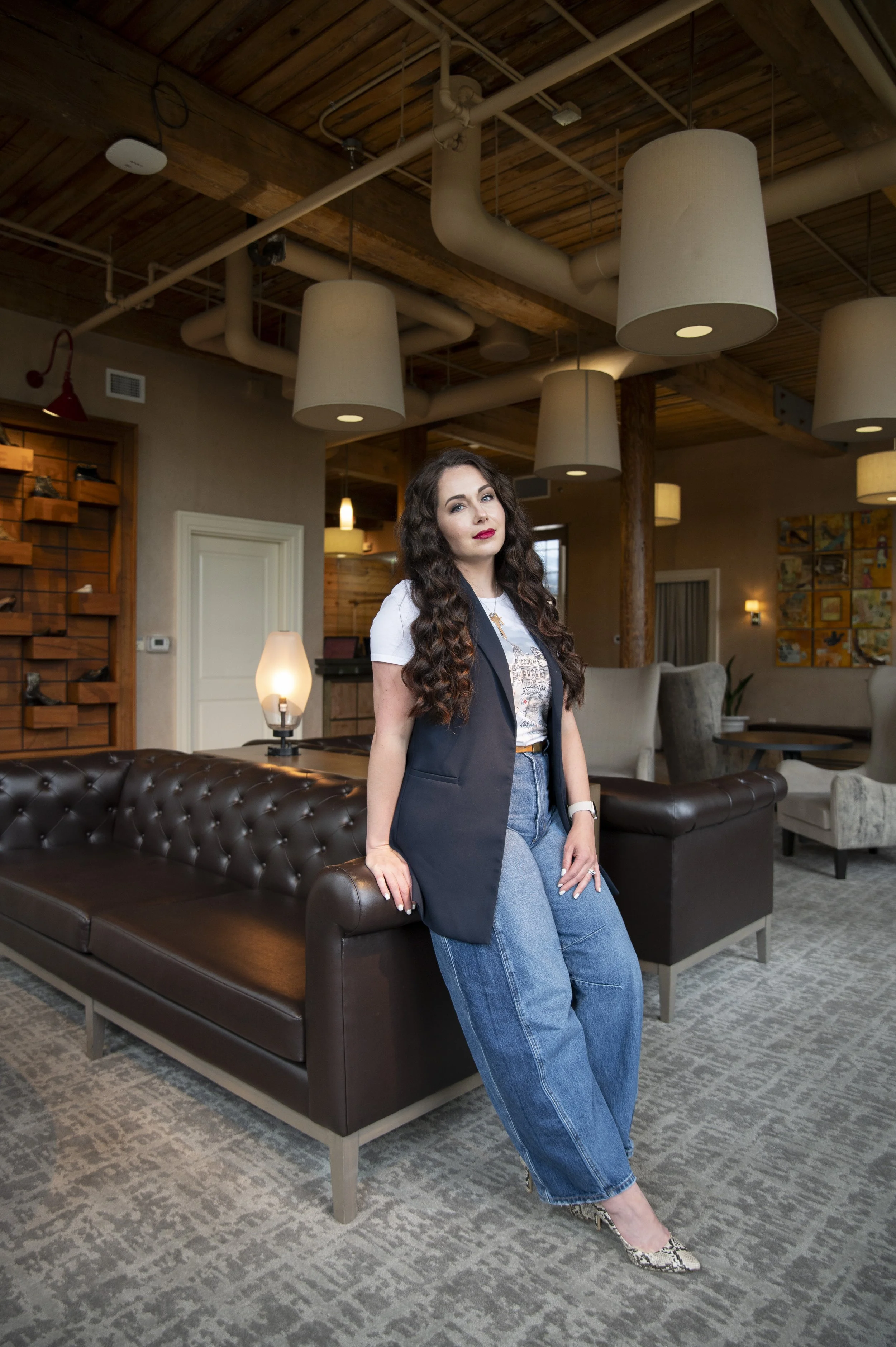 A woman with long wavy dark hair, red lipstick, wearing a white t-shirt, a black blazer, and blue jeans, standing in a cozy lounge area with modern decor, including hanging lamps and wooden furniture.
