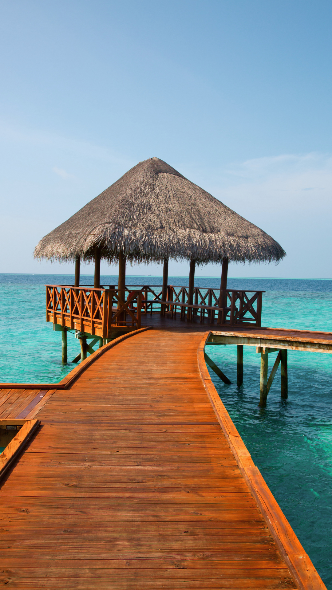 Wooden pathway leading to a thatched-roof hut over clear turquoise water, under a blue sky.