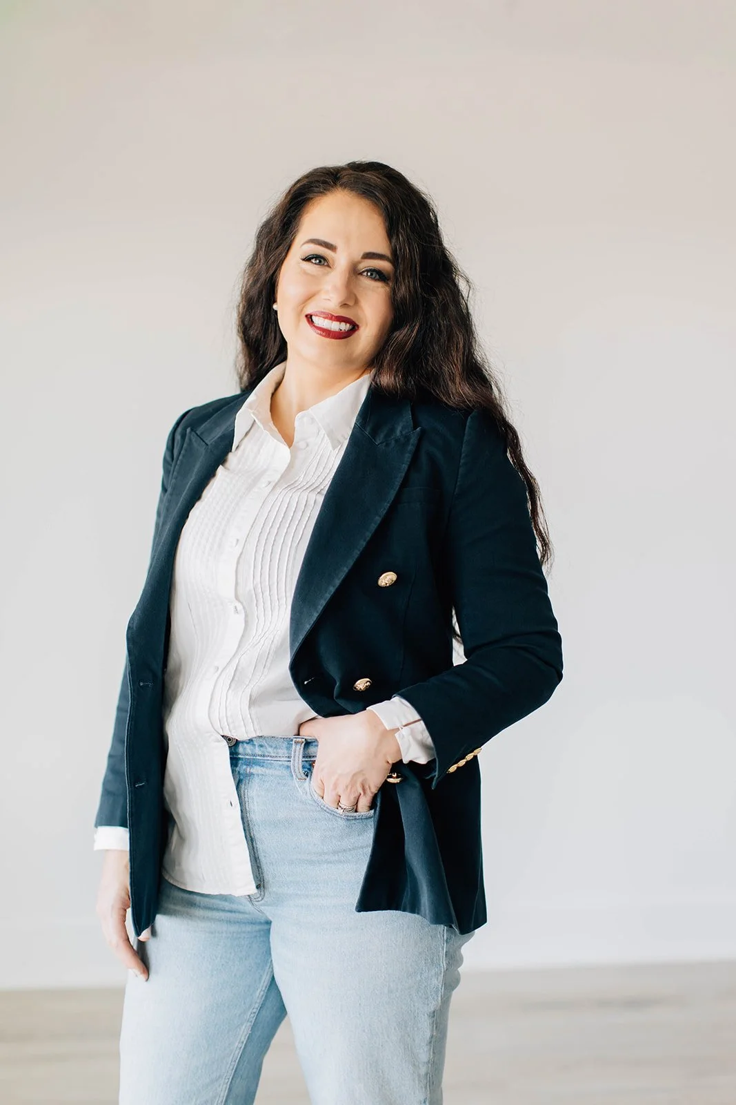 Woman with dark wavy hair wearing a navy blazer, white blouse, and light blue jeans, smiling and posing with her left hand in her pocket against a plain light background.