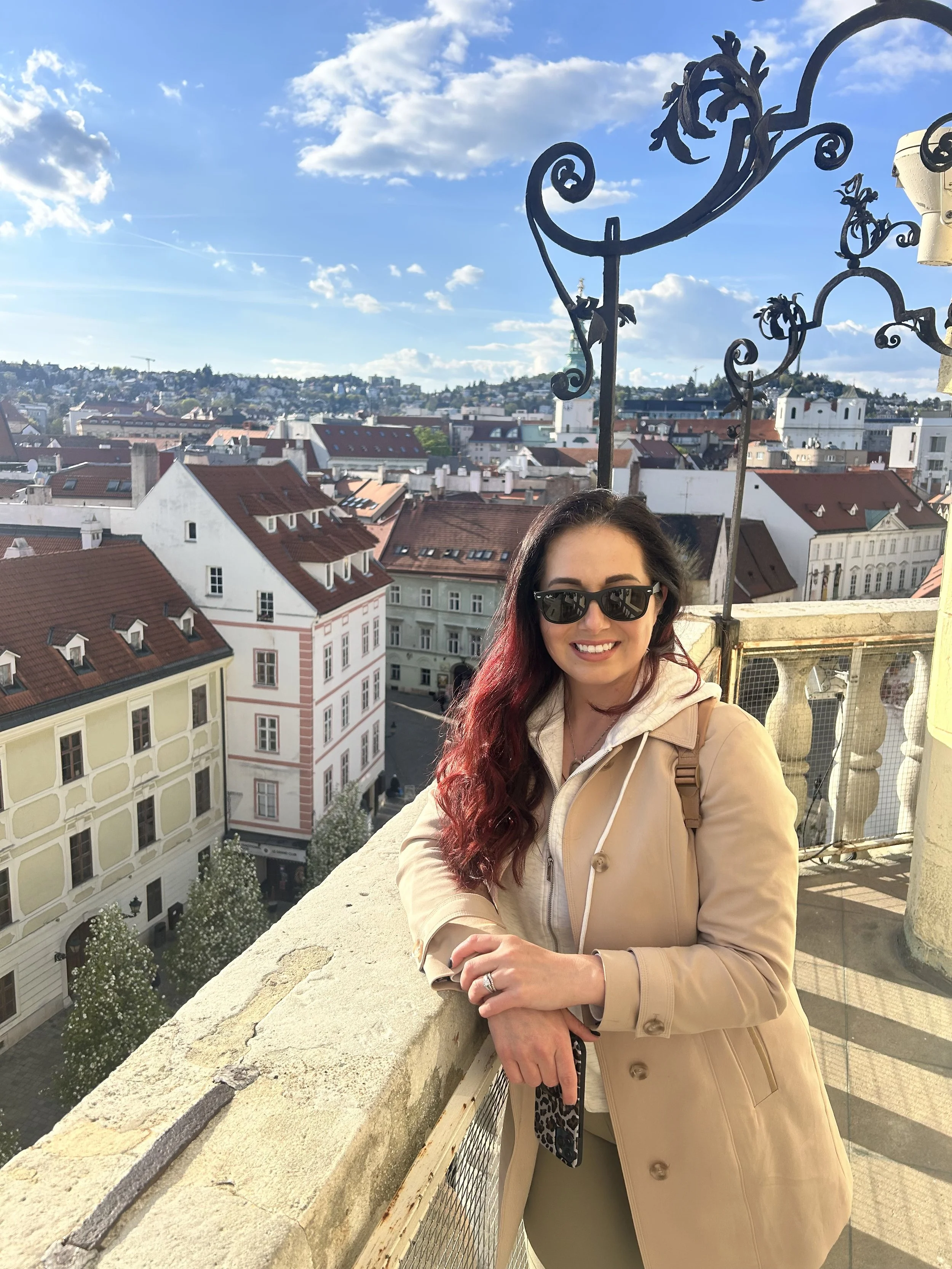 A smiling woman with dark hair and sunglasses taking a selfie on a rooftop, overlooking a city with red-roofed buildings and a blue sky with clouds.