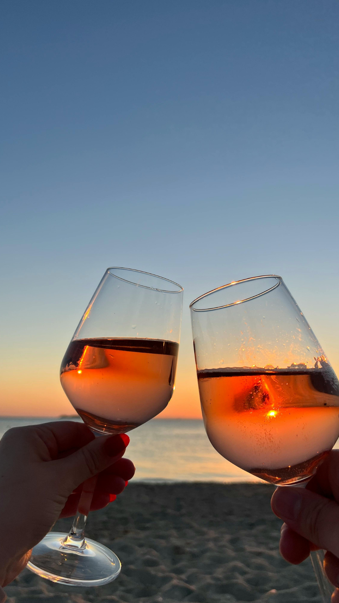 Two glasses of rosé wine being clinked together against a sunset sky at the beach.