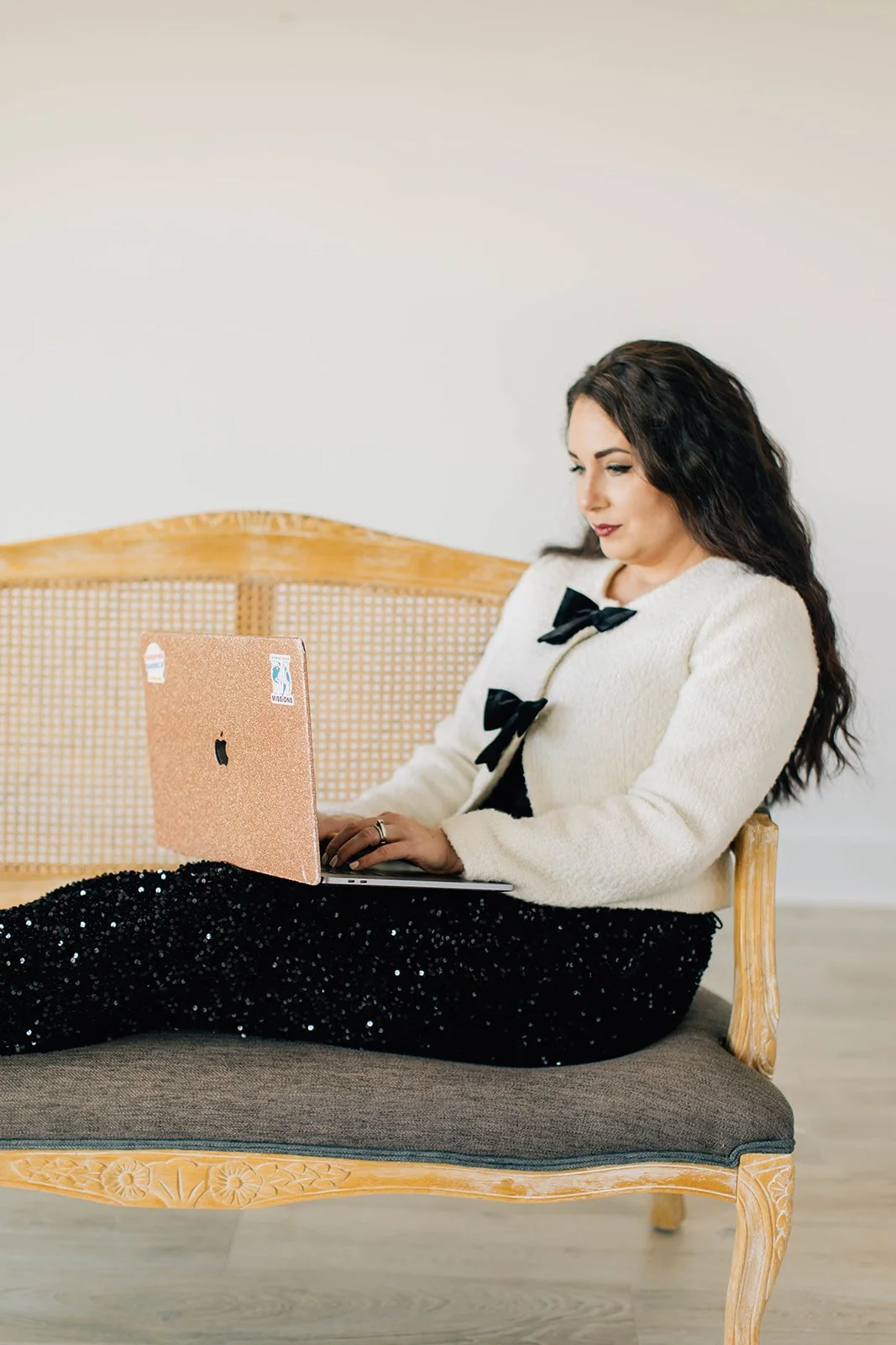 Woman sitting on a vintage wooden bench with a laptop, wearing a white sweater with black bows, and a black sparkly skirt, working indoors.