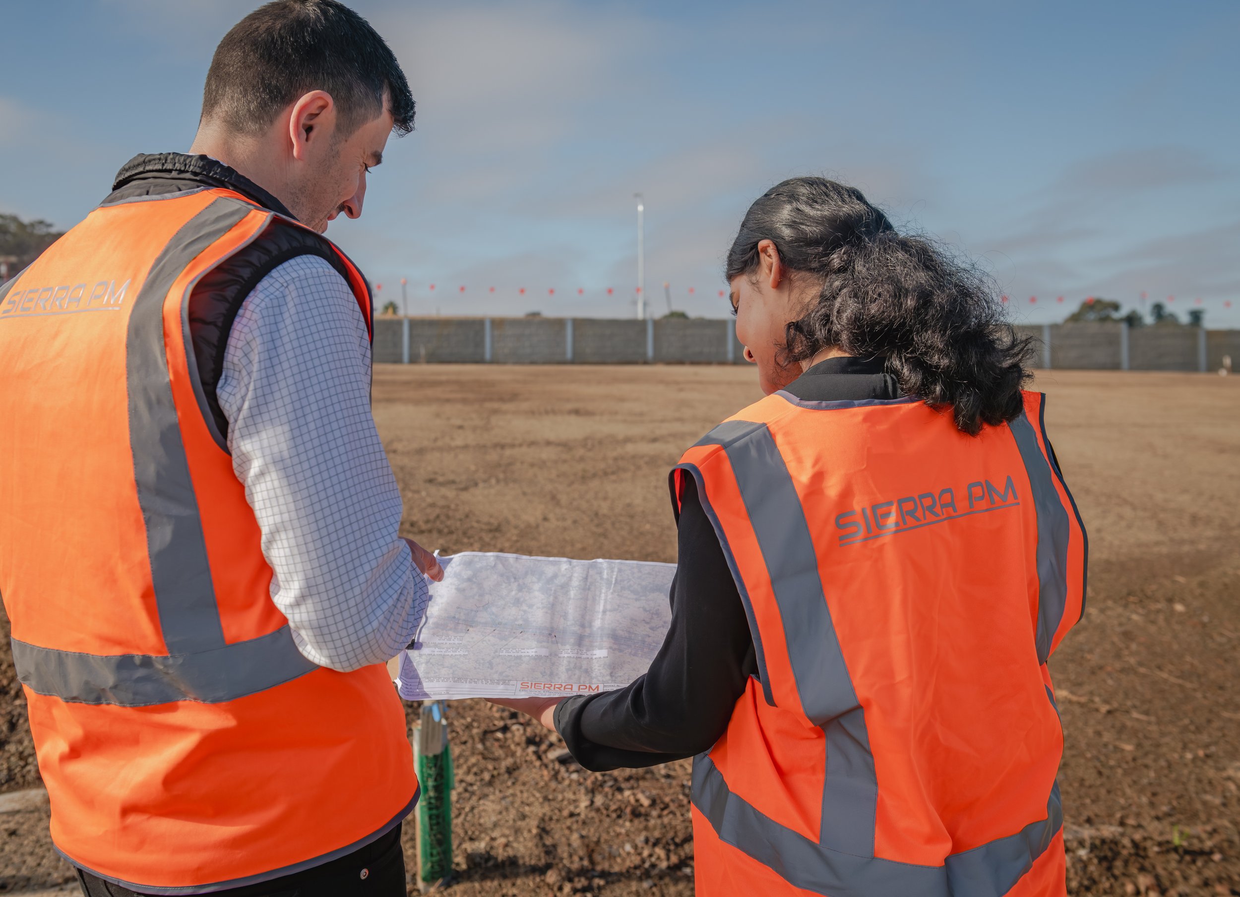 Two people wearing orange safety vests labeled 'SIERRA PM' are examining blueprints on a construction site with bare soil, a fence, and a cloudy sky in the background.
