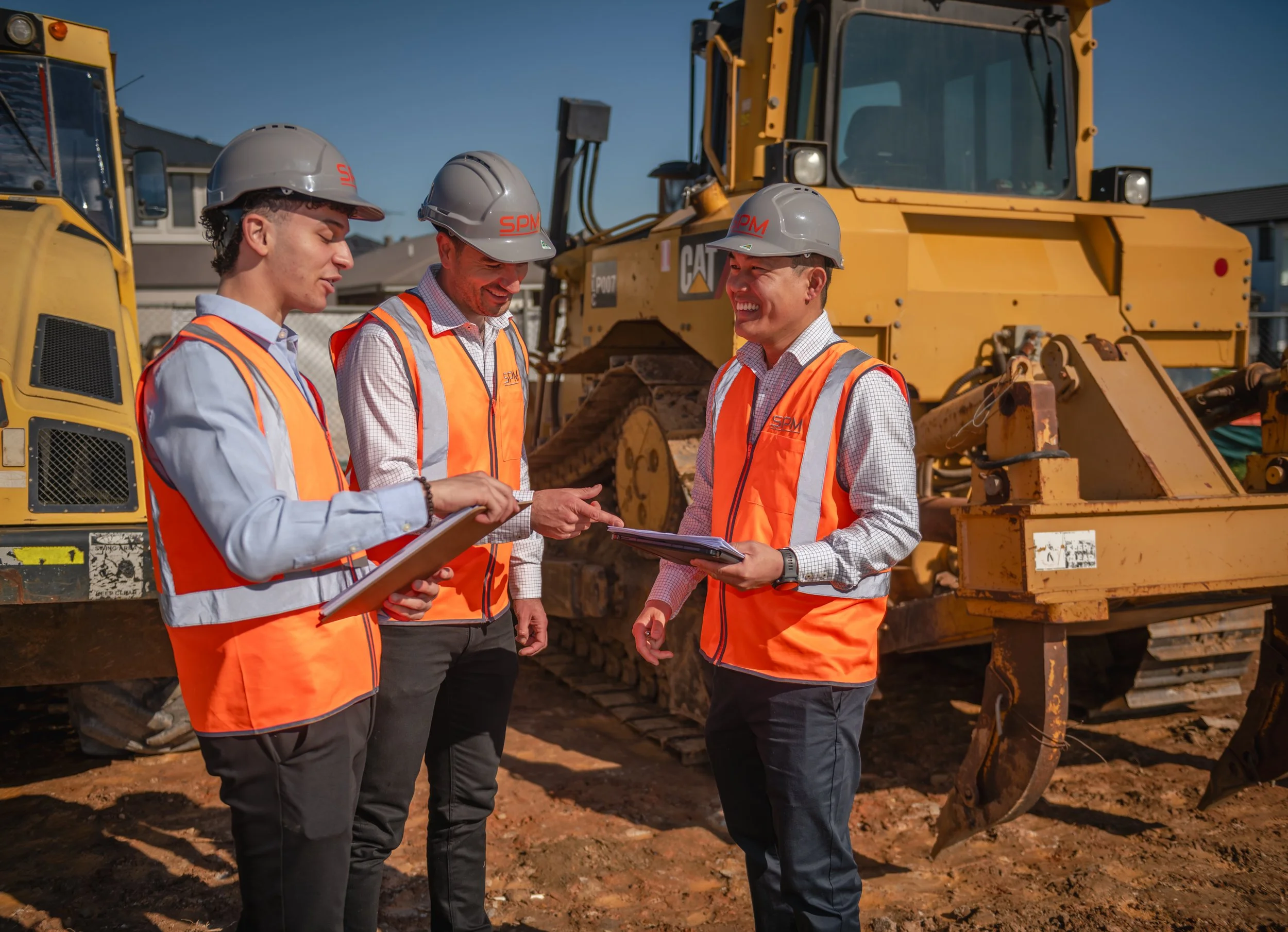 Three construction workers wearing orange safety vests and gray hard hats are standing in front of a large yellow bulldozer, having a discussion outdoors on a construction site, with a building in the background under a clear blue sky.
