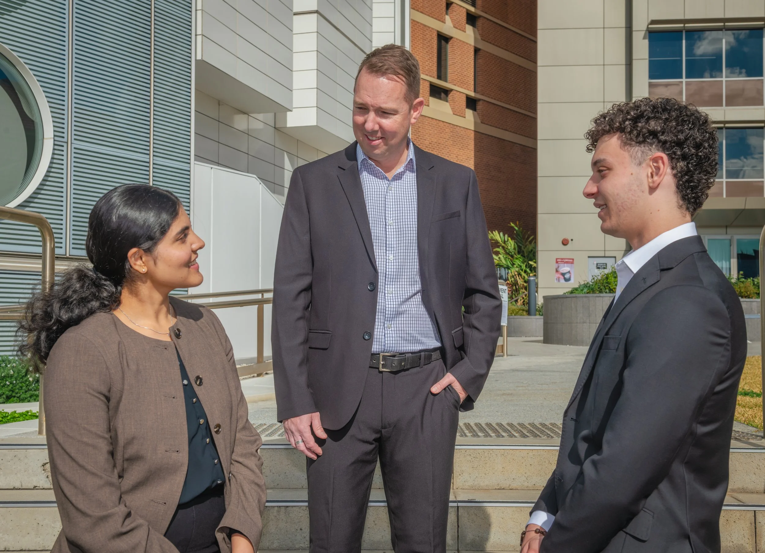 Three people having a conversation outdoors in front of a modern building with glass windows and brick accents. A woman and a man are listening to a second man, who is dressed in a suit.