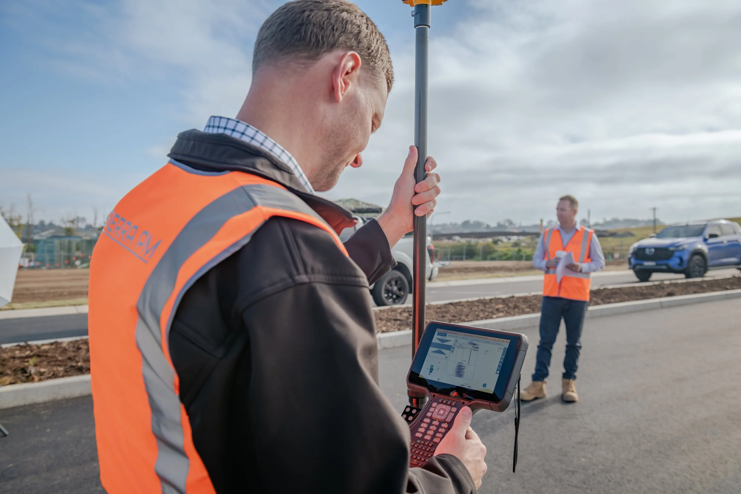 A man wearing an orange safety vest and using a tablet and surveying equipment at an outdoor construction site, with another person in the background and vehicles on the road.