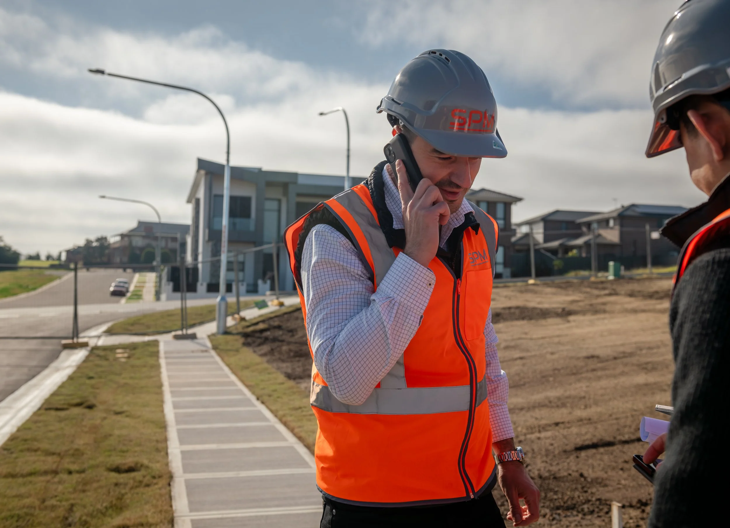 A construction worker in an orange safety vest and gray helmet talking on a cell phone on a residential street under a cloudy sky.
