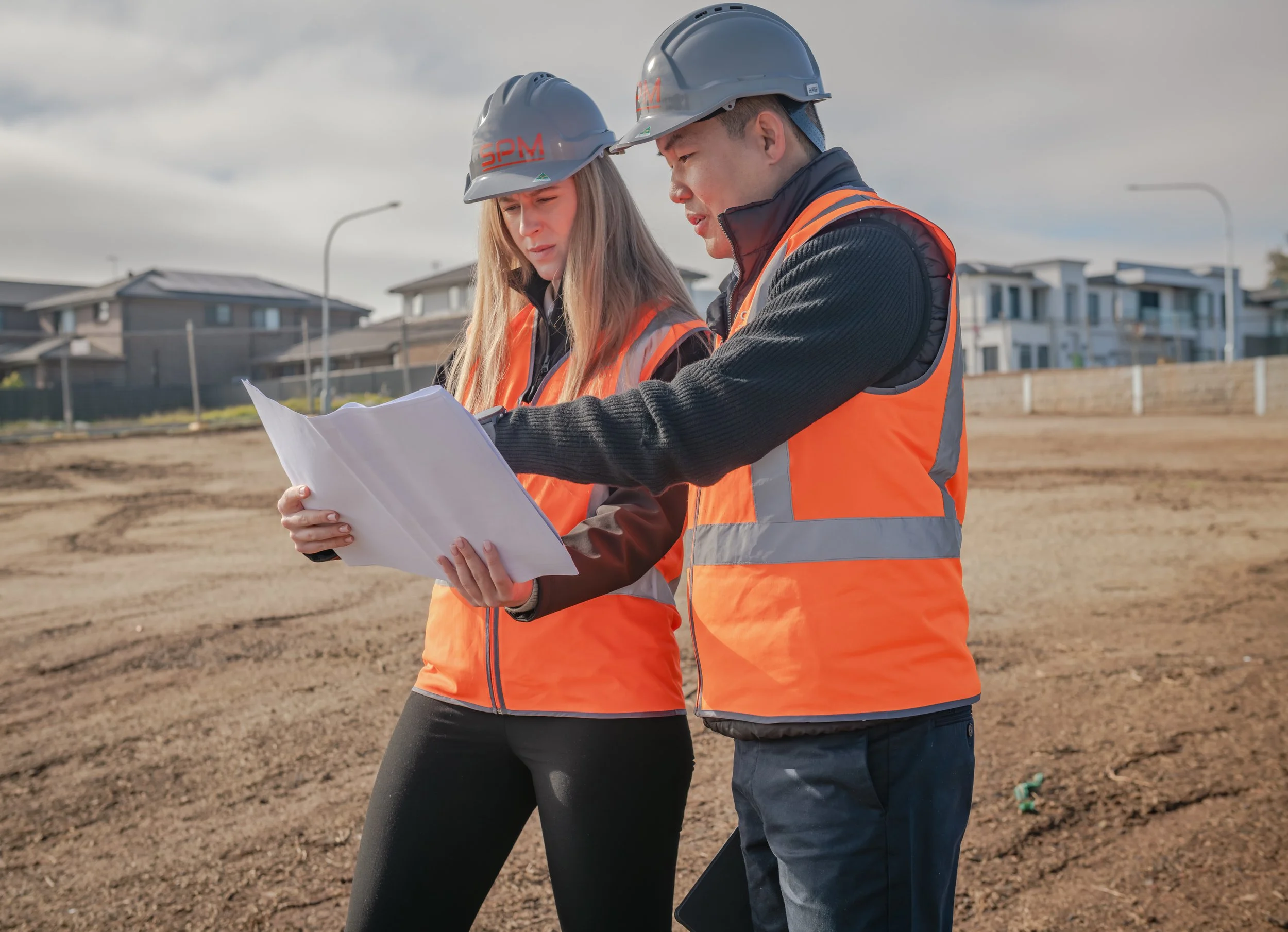 Two construction workers, a woman and a man, wearing safety helmets and orange vests, look at blueprints on a construction site with houses in the background.