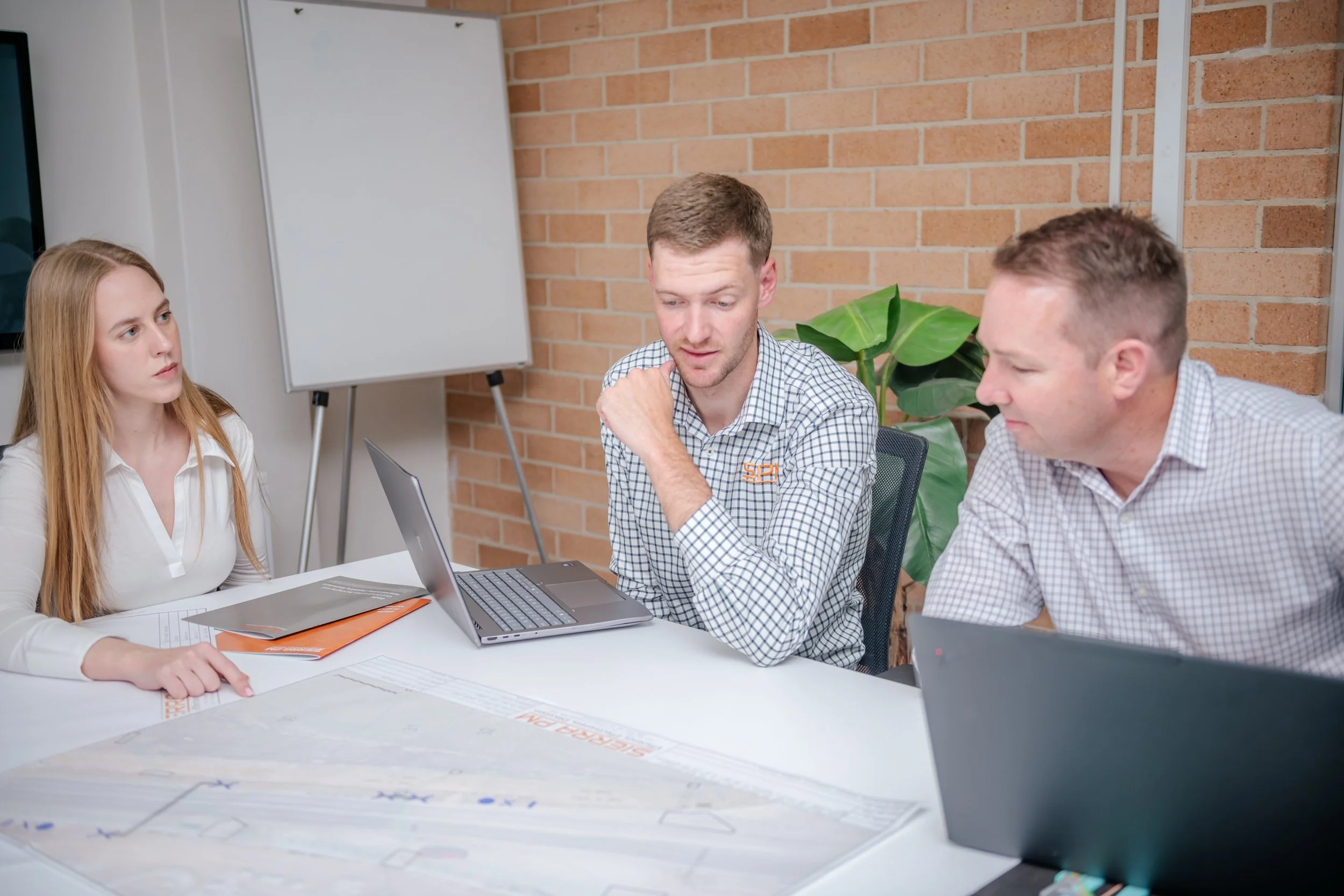 Three people in a meeting room with exposed brick walls, a whiteboard, and a large plant. They are sitting at a table with laptops, papers, and a tablet, engaged in conversation or listening to one person speak.