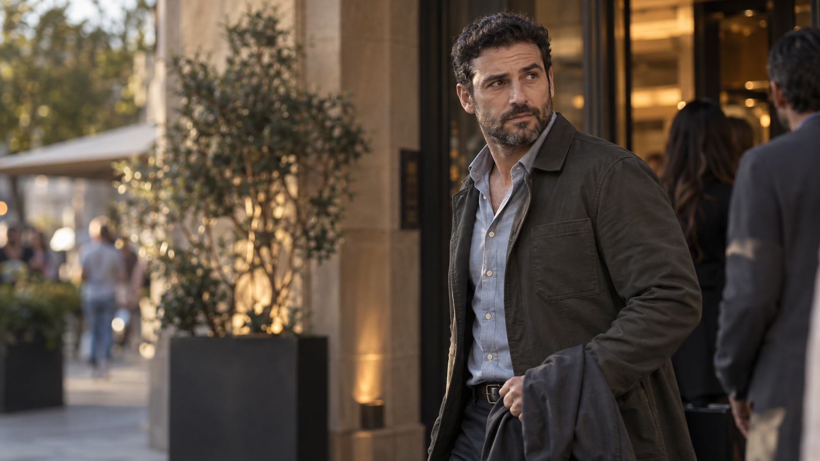 A man with dark, curly hair, a beard, wearing a light blue shirt and a dark jacket, looking to the side outside a building with large windows and people in the background.