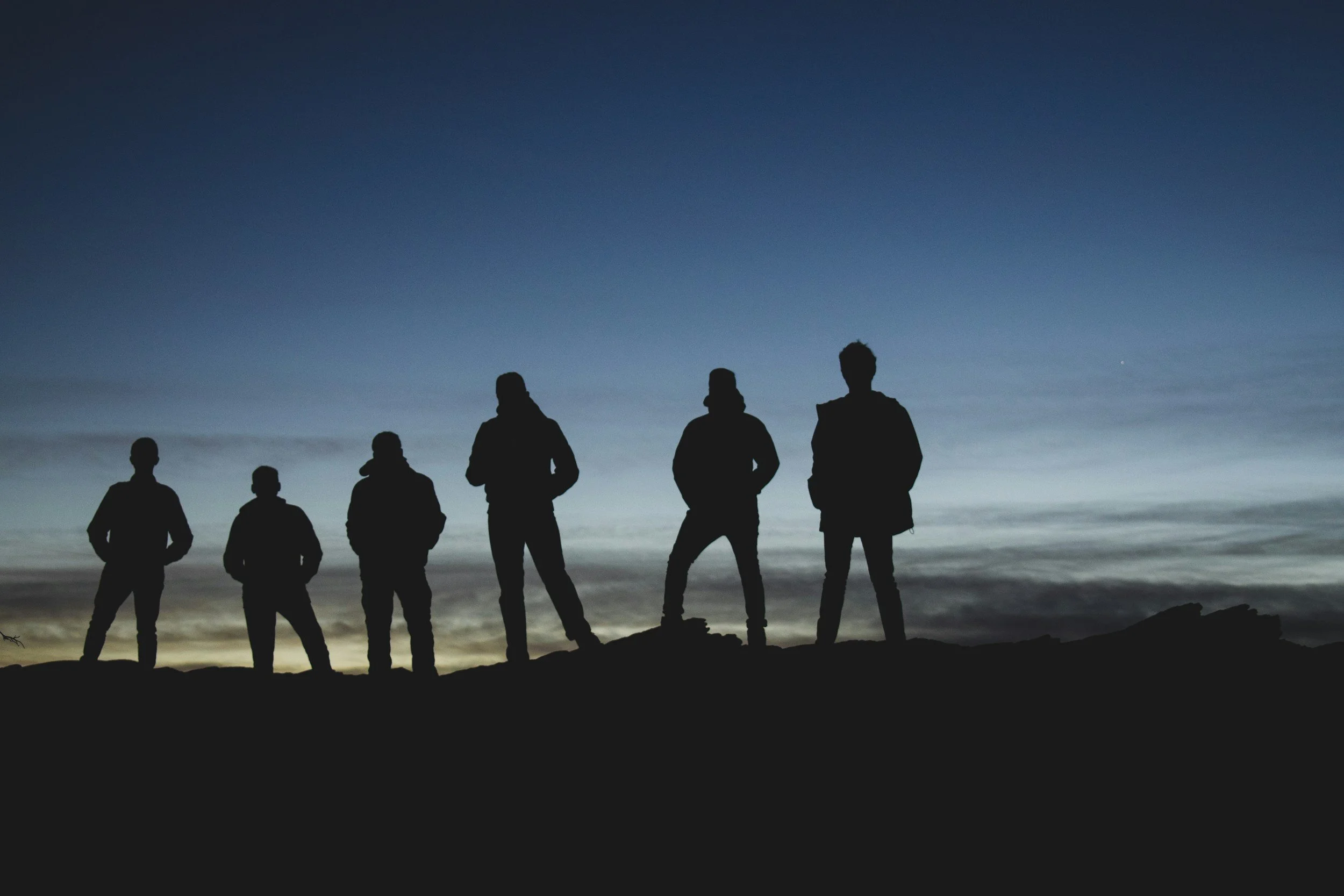 Silhouettes of seven people standing on a hill at sunset or sunrise with a dark blue sky and a faint horizon line.