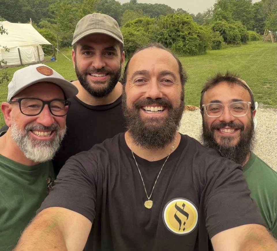 Four men smiling for a selfie outdoors in a grassy area with trees, a tent, and playground equipment in the background.
