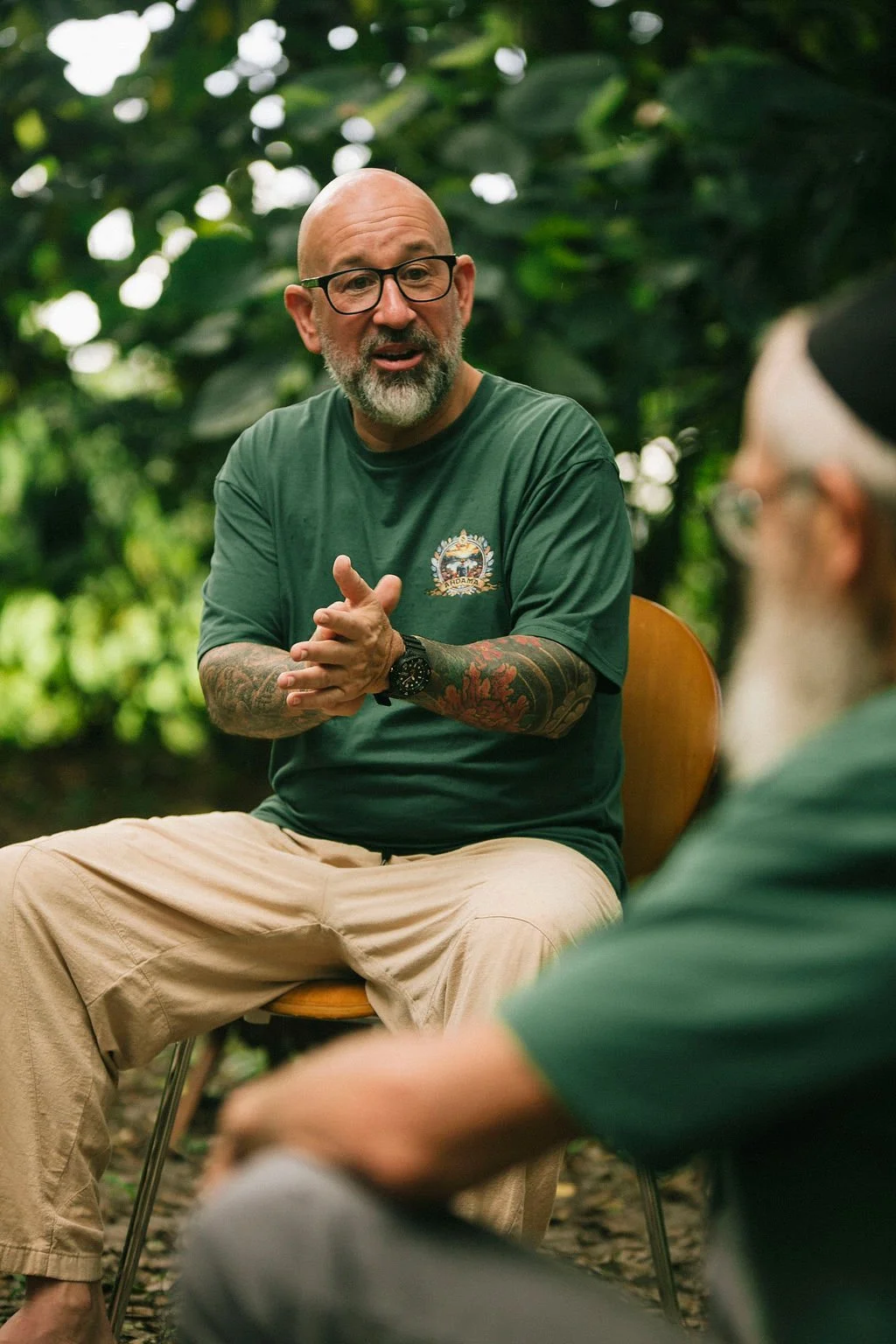 A bald man with glasses, tattoos on his arms, and a beard, is sitting outdoors in a lush, green environment with trees. He is wearing a green t-shirt, beige pants, and a wristwatch, and appears to be engaged in a conversation with another person.