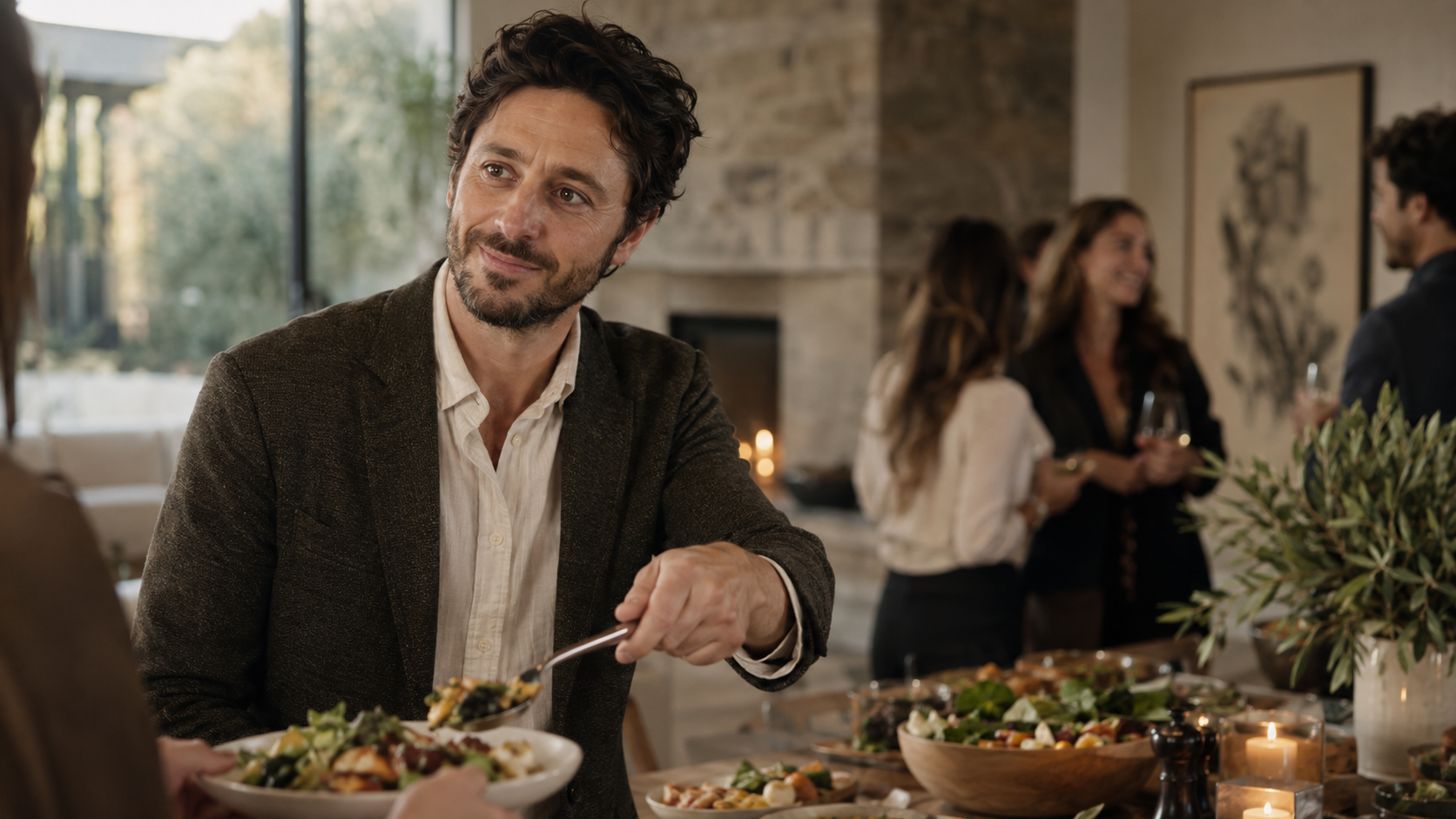 A man with dark wavy hair and a beard, wearing a brown blazer and white shirt, is smiling while holding a plate of food at a social gathering in a well-lit room with large windows, other guests in the background, and a buffet table with salads and candles.