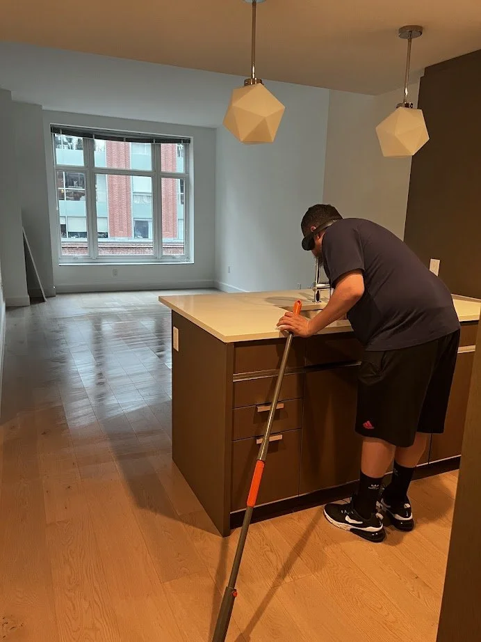 Person mopping a wooden floor in a modern kitchen with pendant lights and large window.