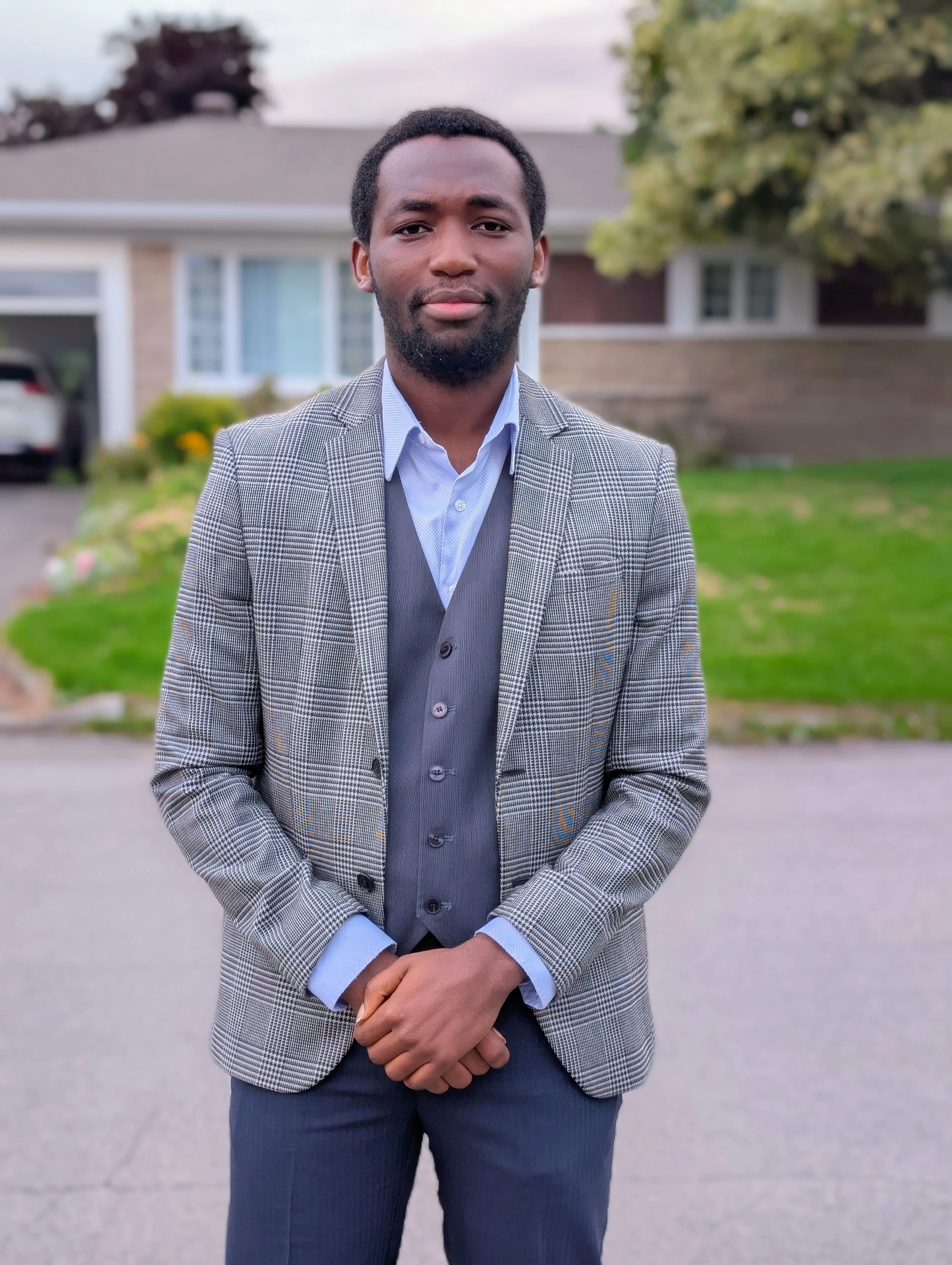 A man stands outdoors in front of a house, wearing a gray checked blazer over a light blue shirt and dark pants, with his hands clasped in front of him, during the daytime with an overcast sky.