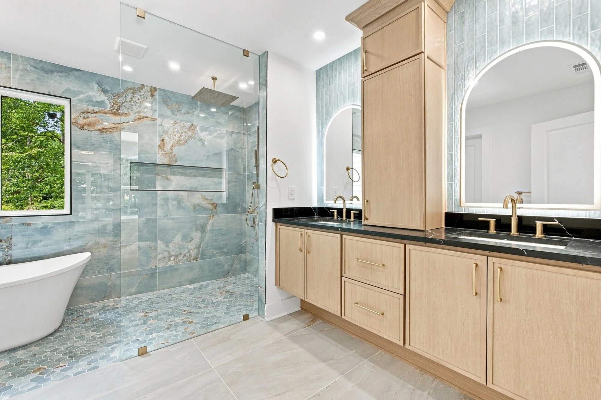 Modern bathroom with a walk-in shower featuring blue and gold marble tiles, a large window with greenery outside, a freestanding bathtub, a double vanity with black countertop, and light wood cabinets with gold handles. There are two illuminated mirrors above the vanity, gold fixtures, and a terrazzo tile floor.