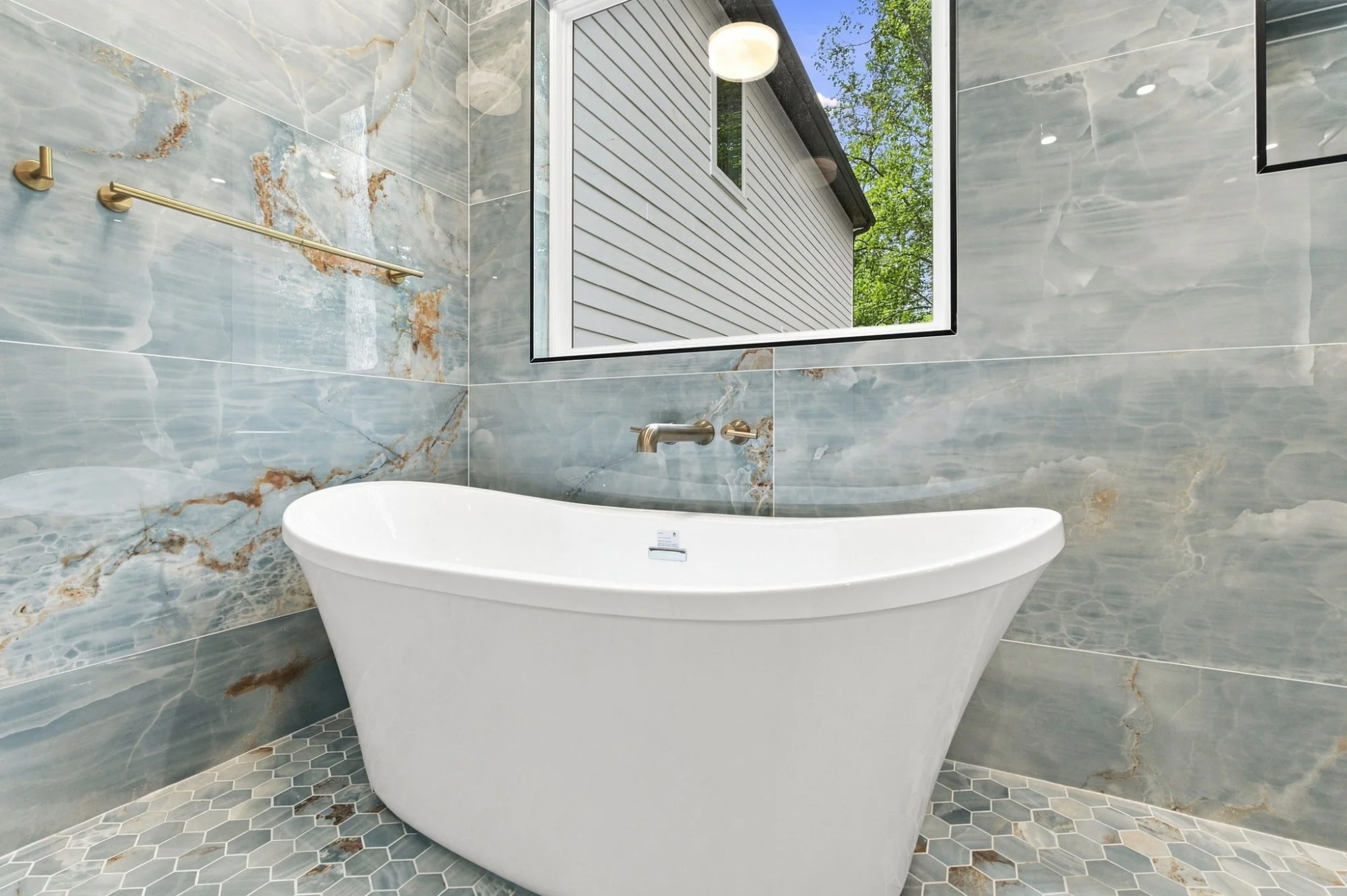 Modern bathroom with a freestanding bathtub, marble tile walls, a large window showing a house exterior and trees, and a hexagonal tile floor.