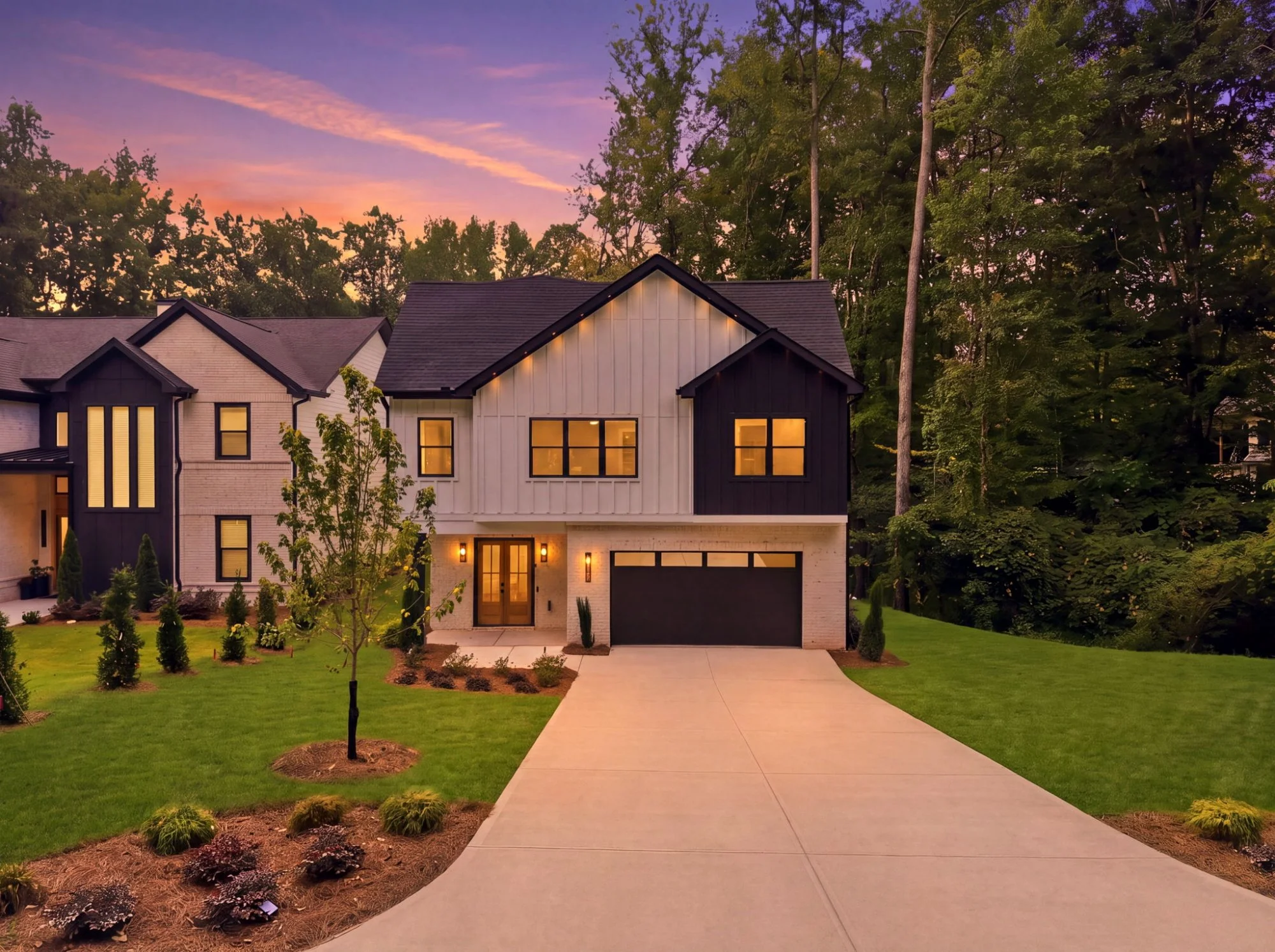 Modern house with black and white exterior, large windows glowing with interior lights, surrounded by a manicured lawn and small trees, with a sidewalk leading to the front door, against a colorful sunset sky with tall trees in the background.