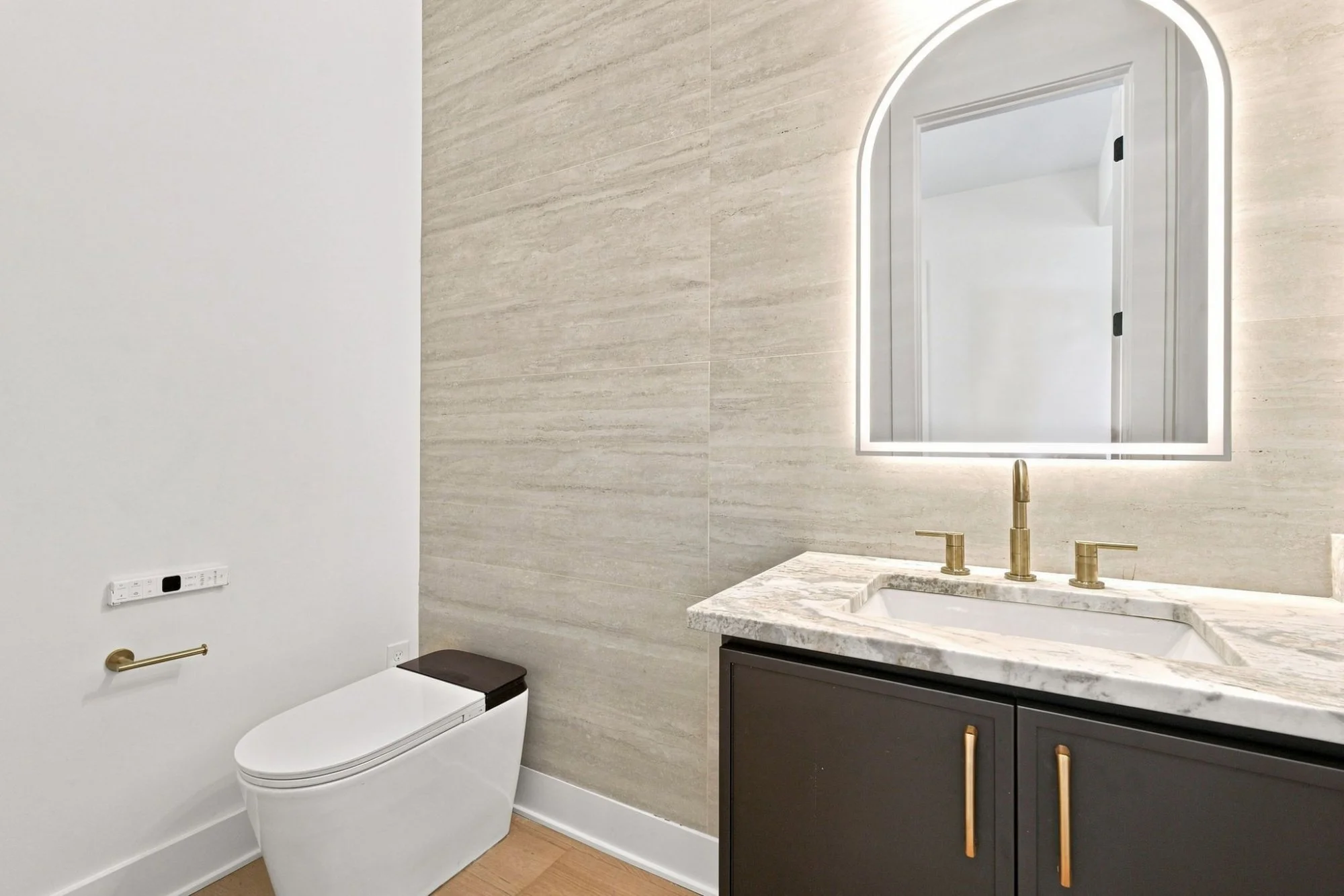 Modern bathroom with beige marble countertop, black vanity cabinet with gold handles, illuminated arched mirror, gold faucet, white toilet, white wall with electrical outlets, and strip lighting around the mirror.