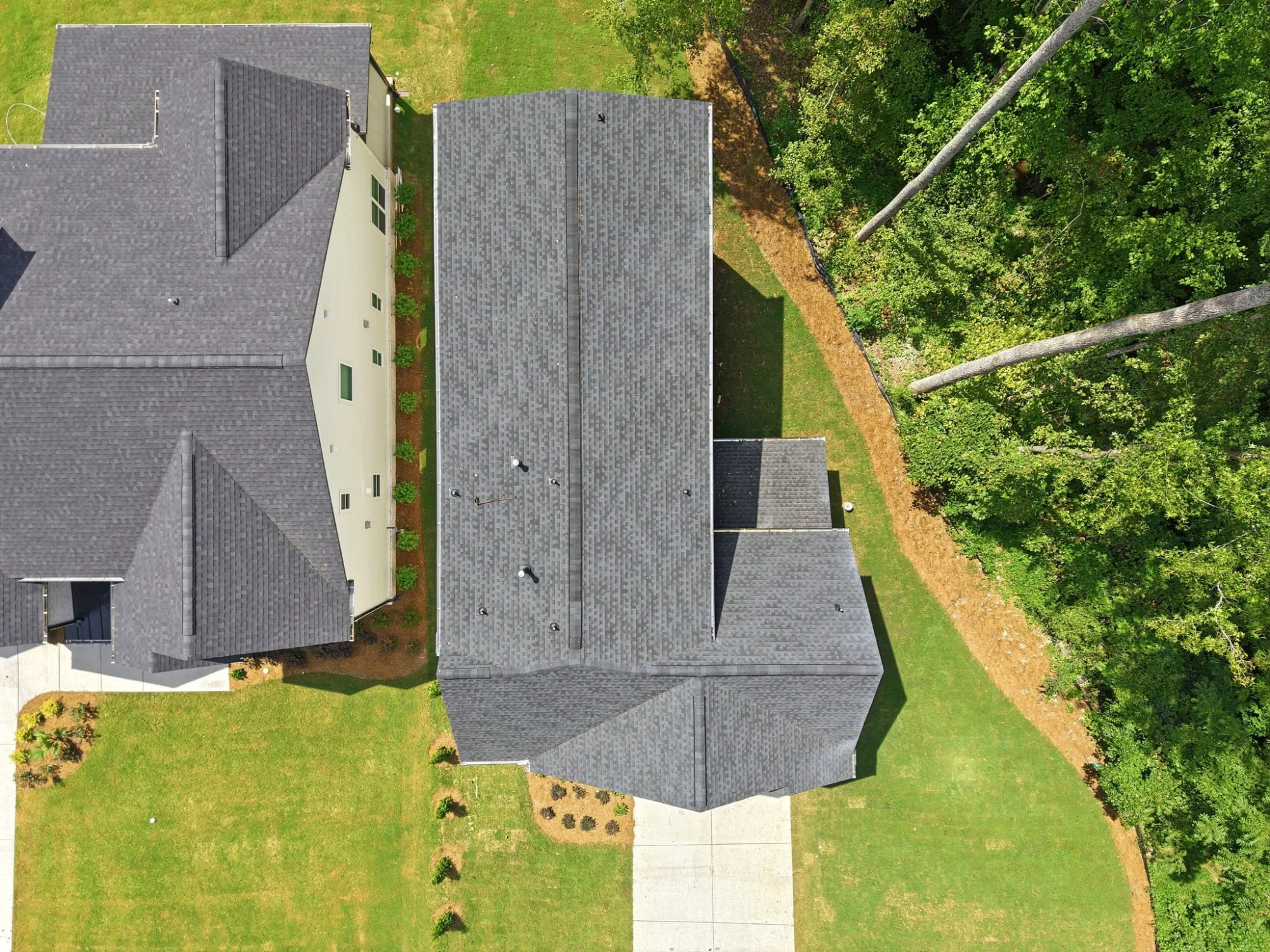 An aerial view of a residential house with multiple dark gray shingled roofs, a small yard with a concrete walkway, lush green trees, and bushes along the yard's perimeter.