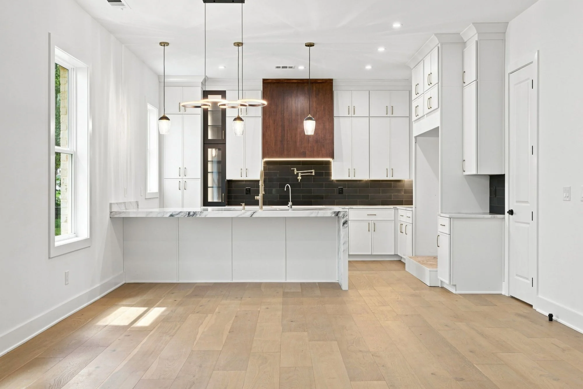 Empty modern kitchen with white cabinets, black backsplash, marble countertop, wooden floors, and large windows.