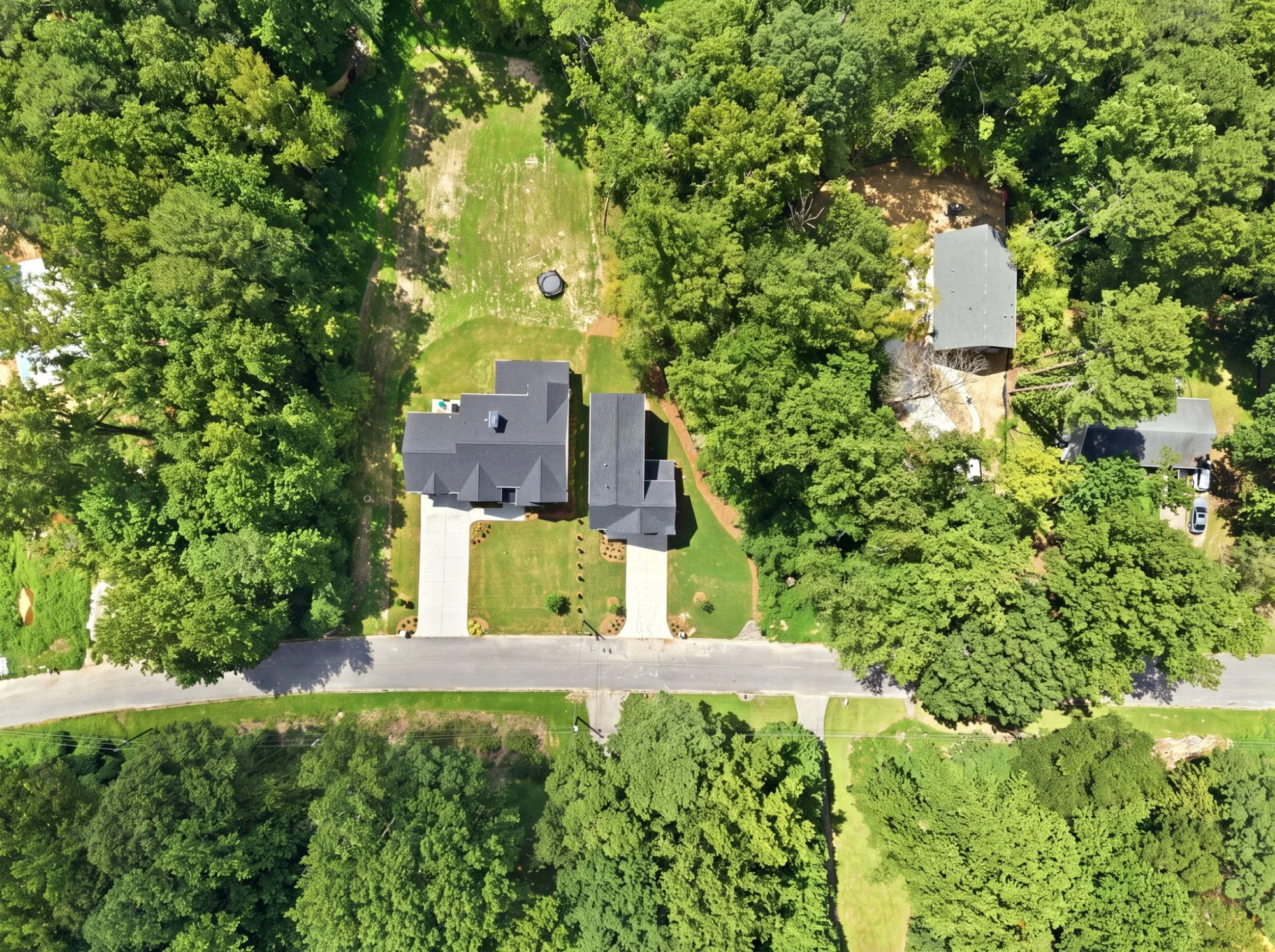 Aerial view of a residential neighborhood with a house, large trees, a lawn, a trampoline, and a driveway, surrounded by greenery.
