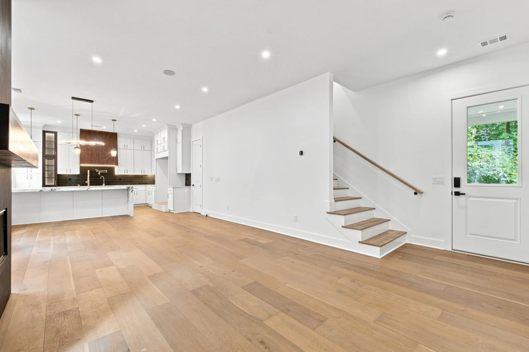 Interior of a modern, bright living space with wooden flooring, white walls, a staircase with wooden steps, large window showing greenery outside, and a kitchen area with white cabinets and black backsplash.