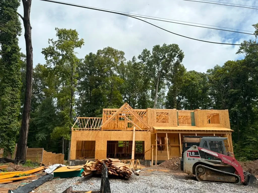 Construction site with a partially built two-story wooden house, surrounded by trees, with construction equipment and materials on the ground.