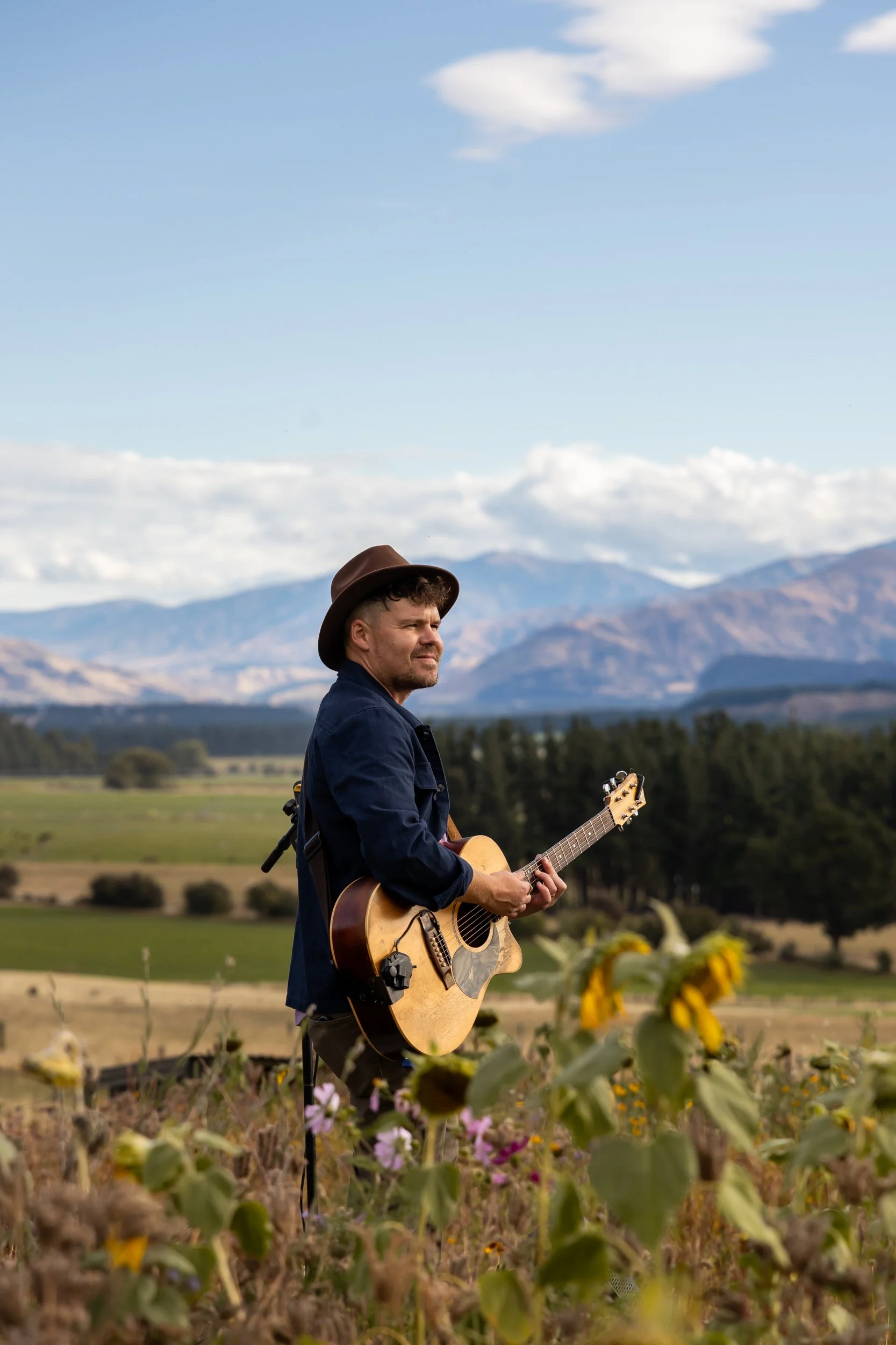 Artist Josh Leys performing holding his guitar at the flower field entrance to Lake Hawea Station