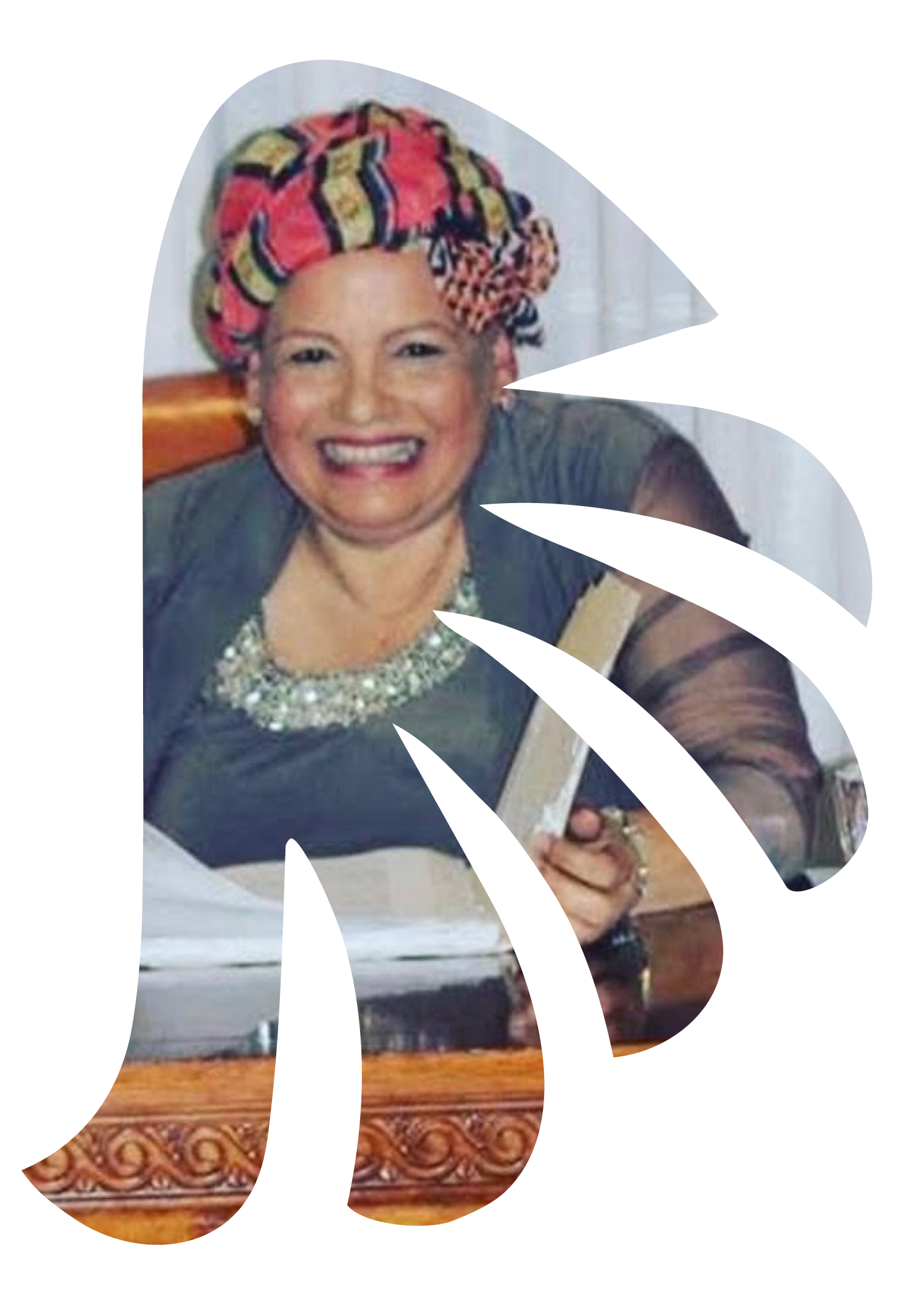 A smiling woman with makeup and a colorful headwrap sitting at a table with a white tablecloth and a wooden base, in a room with a white door.