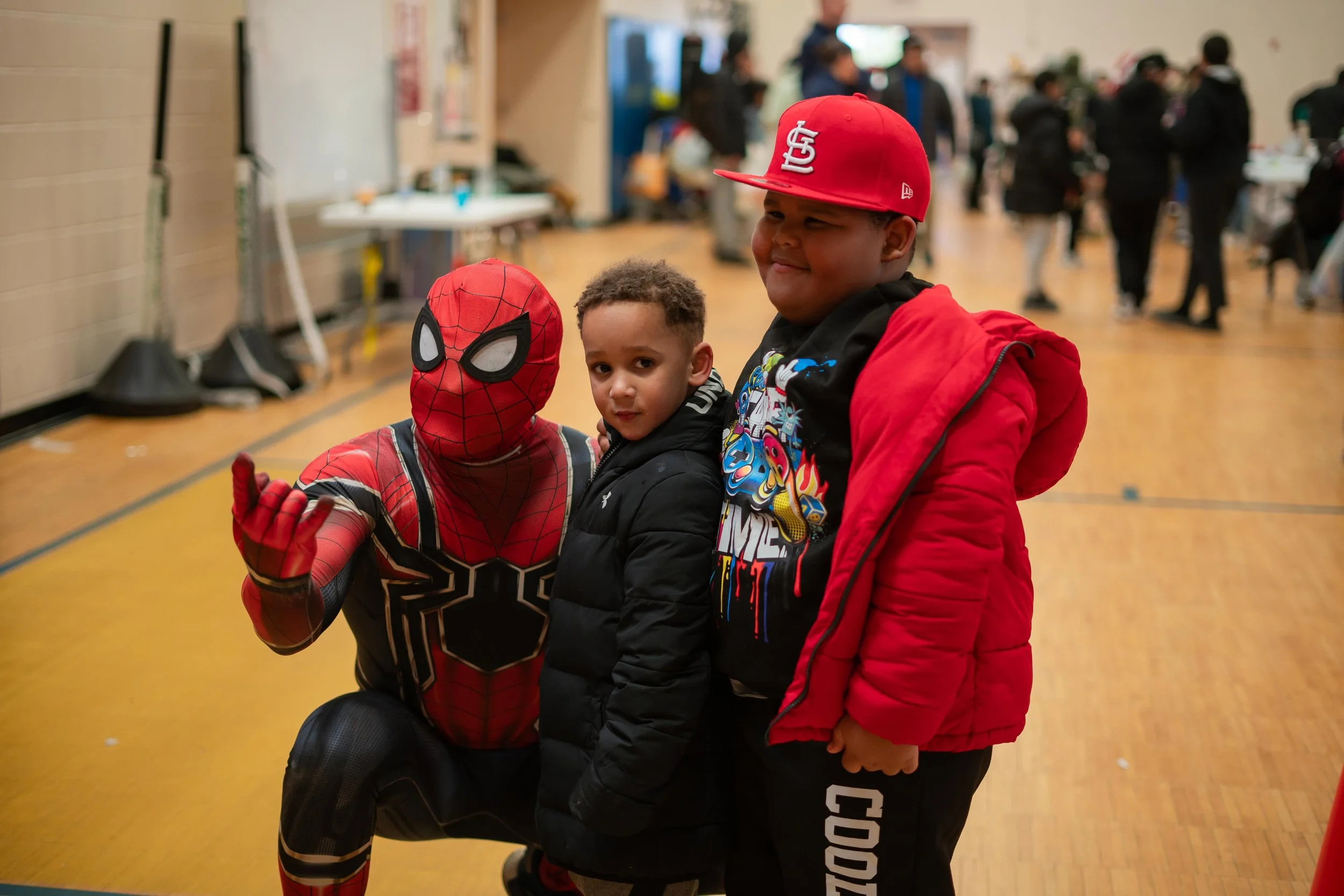 Three young boys dressed as Spider-Man, a young boy in casual clothing with a black jacket and another in a red hoodie and cap, standing in a gymnasium with blurred background of other children and adults.
