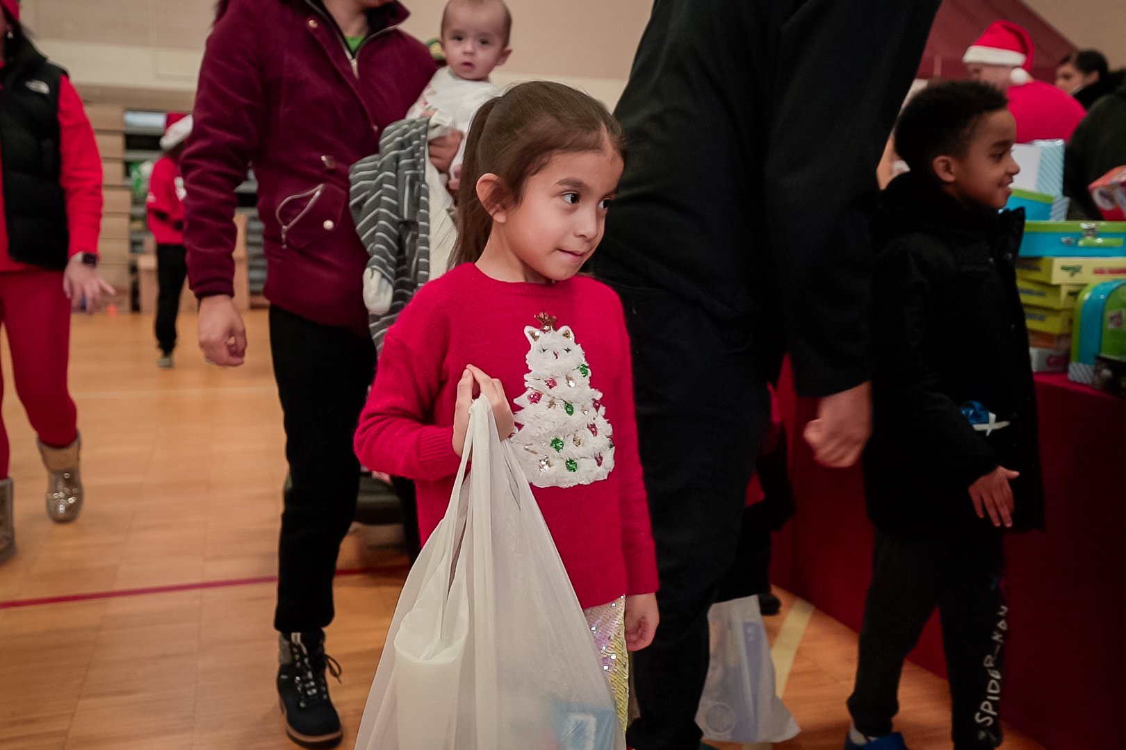 A young girl wearing a red Christmas sweater with a white Christmas tree design, carrying a white shopping bag, waiting in line at a festive event, with several children and adults in the background.
