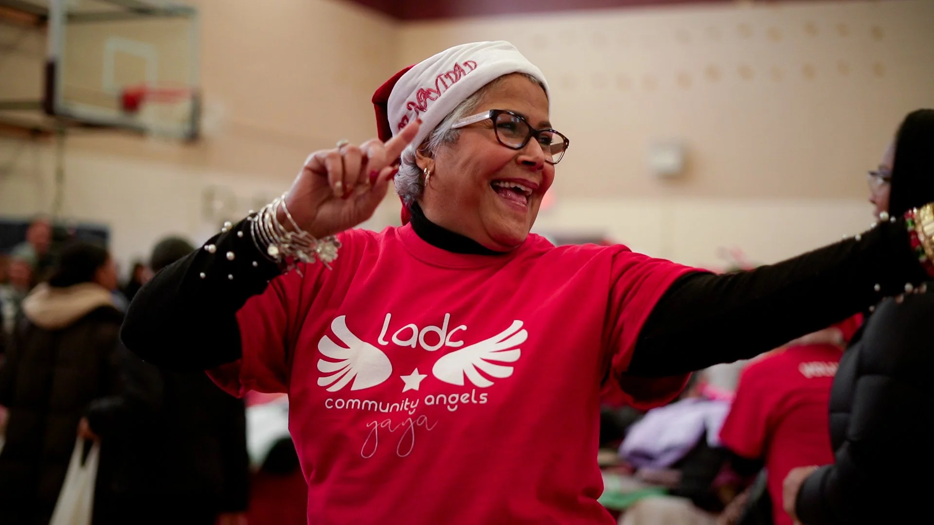 Woman smiling and pointing with her right hand, wearing a red T-shirt with a logo and the words "community angels yoga," glasses, earrings, and a red and white Santa hat. She is indoors at a community event with people in the background.