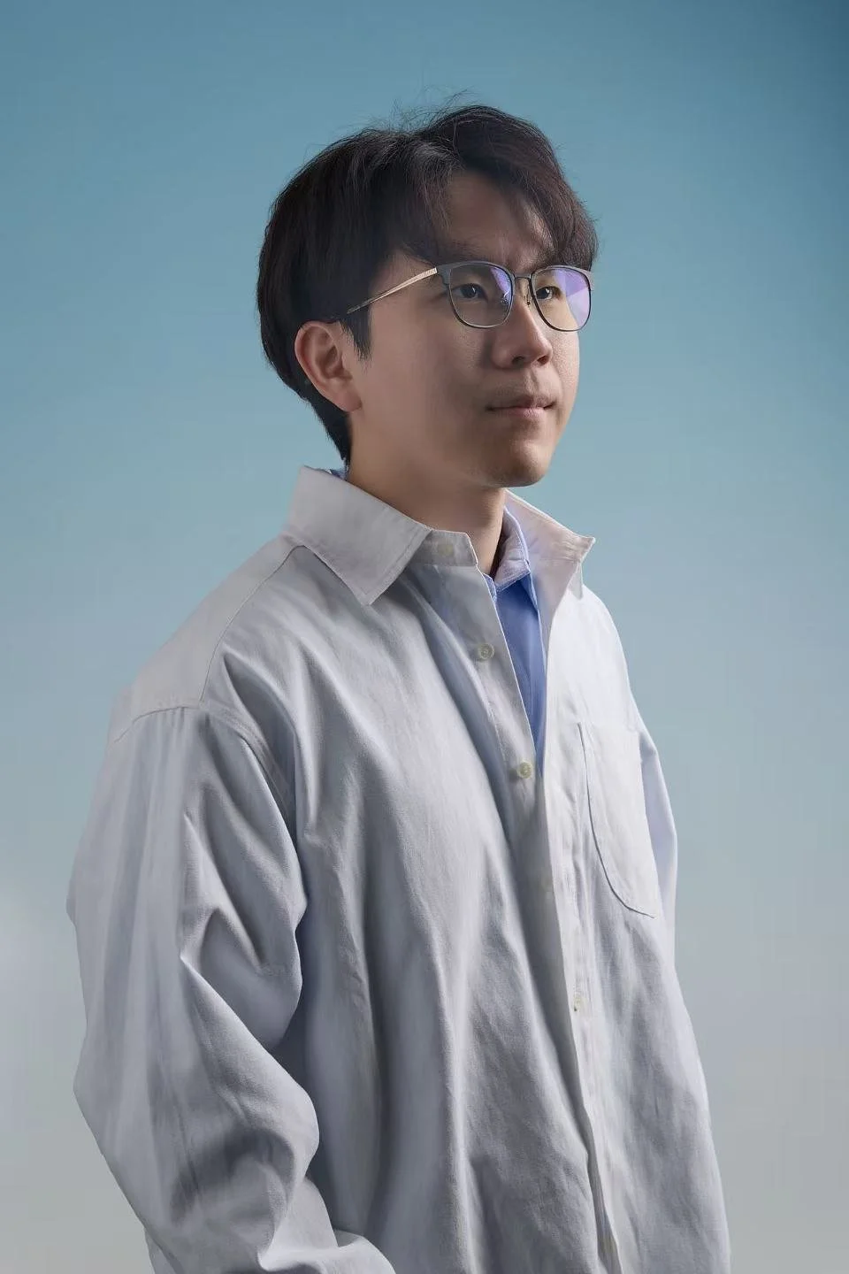 A young Asian man with black hair and glasses, wearing a light gray button-up shirt over a blue collared shirt, posed against a light blue background.