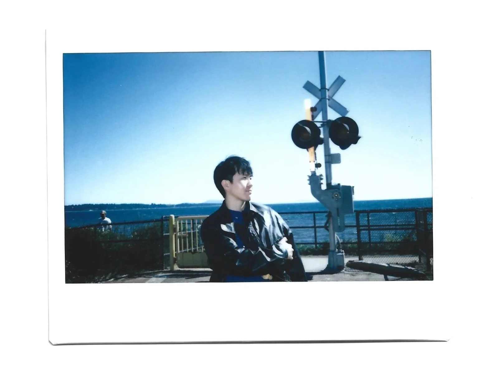A young man with dark hair and a leather jacket stands near a railroad crossing signal, with arms crossed and a slight smile, overlooking a body of water under a clear blue sky.