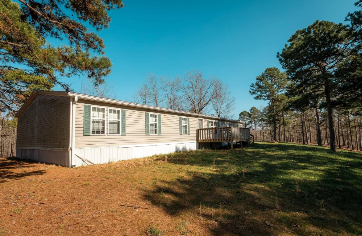 Single-story beige manufactured home with green shutters on windows, located in a wooded area with trees, grass, and a small deck outward from the house under a bright blue sky.