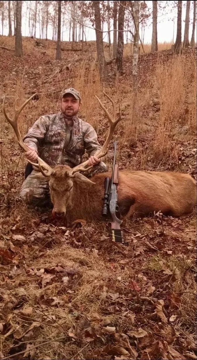 A man in camouflage clothing kneels next to a large freshly killed deer with antlers, holding the antlers. The deer is laying on the ground in a wooded area with trees and brown fallen leaves.