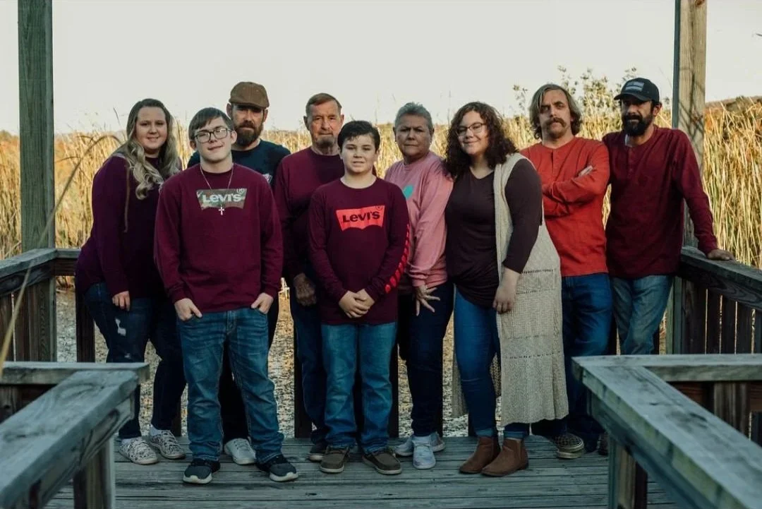 Family group photo on a wooden deck outdoors with tall grass and trees in the background.