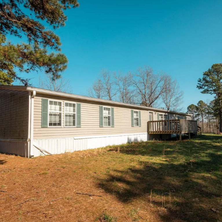 Single-story mobile home with beige siding, white trim, and green shutters, surrounded by trees and grass on a sunny day.